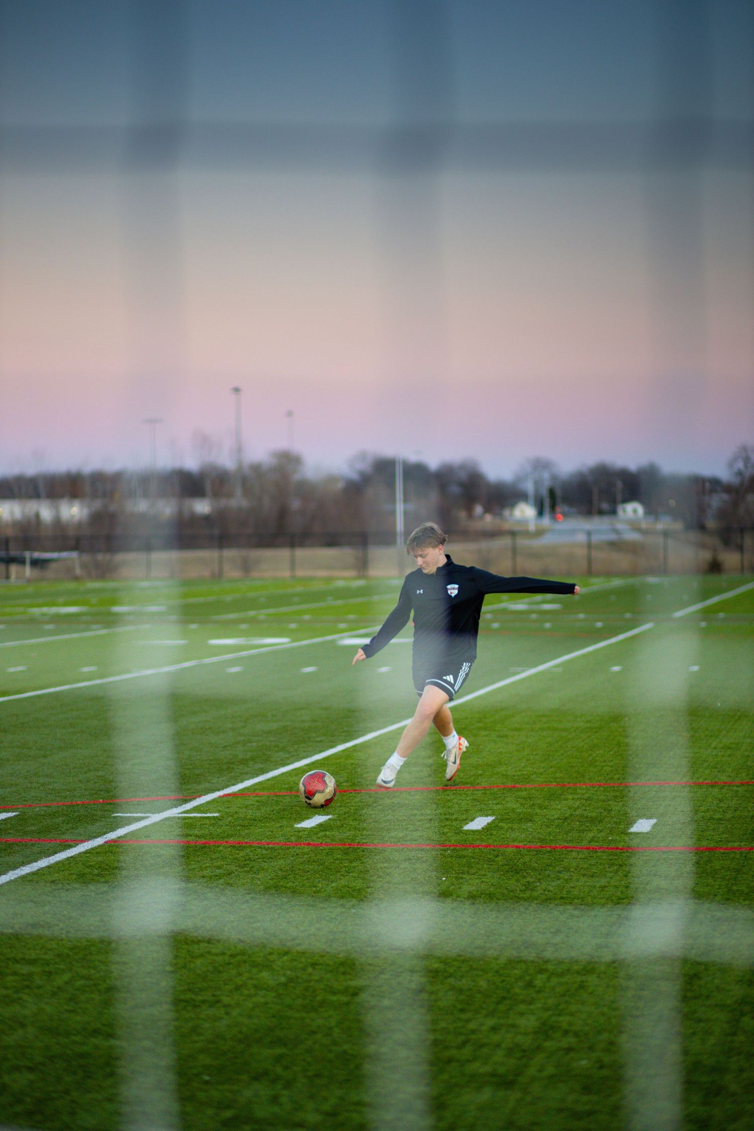 Young soccer player kicking ball on green turf field at sunset, photographed through goal netting.