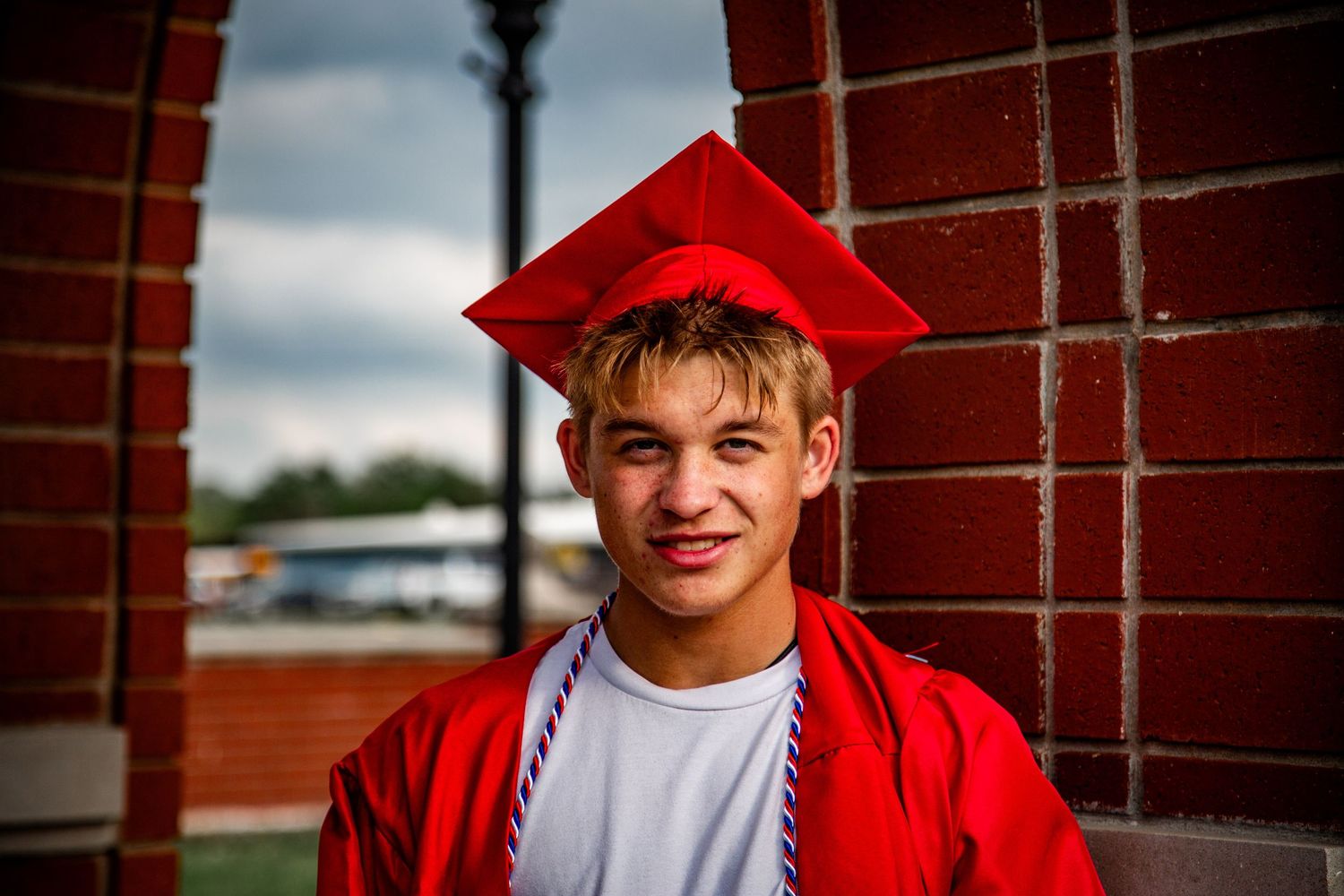 Smiling teenage boy in red graduation cap and gown against brick wall.
