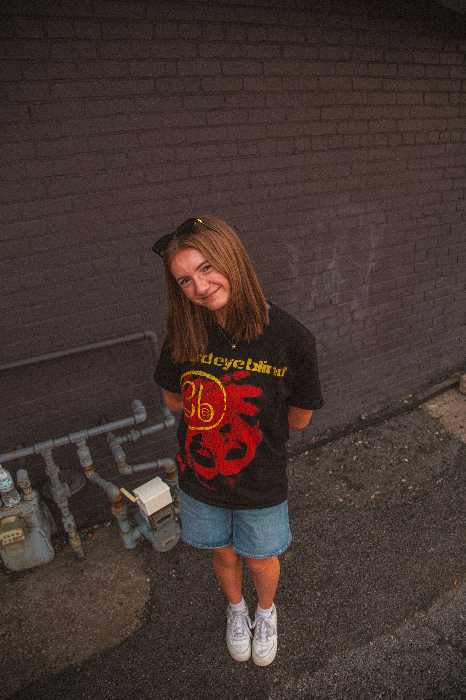 Young woman in a Third Eye Blind band tee and denim shorts smiling against a brick wall outdoors.