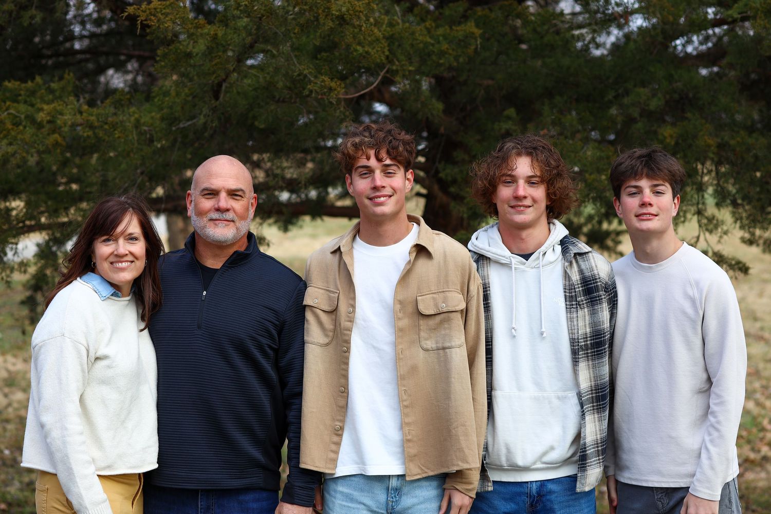 A smiling family of five stands together outdoors with green trees in the background.