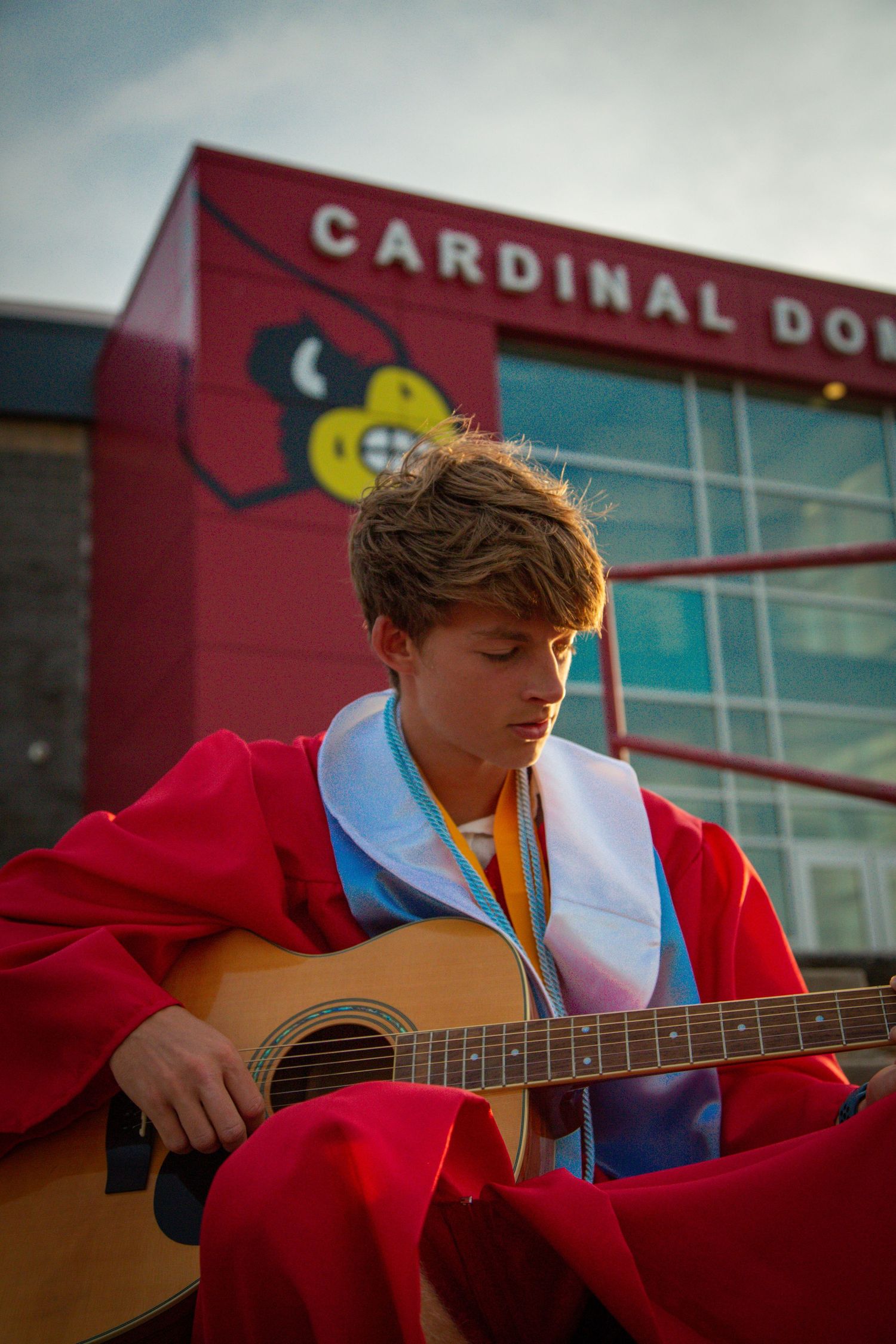 High school graduate in red gown plays acoustic guitar outside Cardinal Dome building.