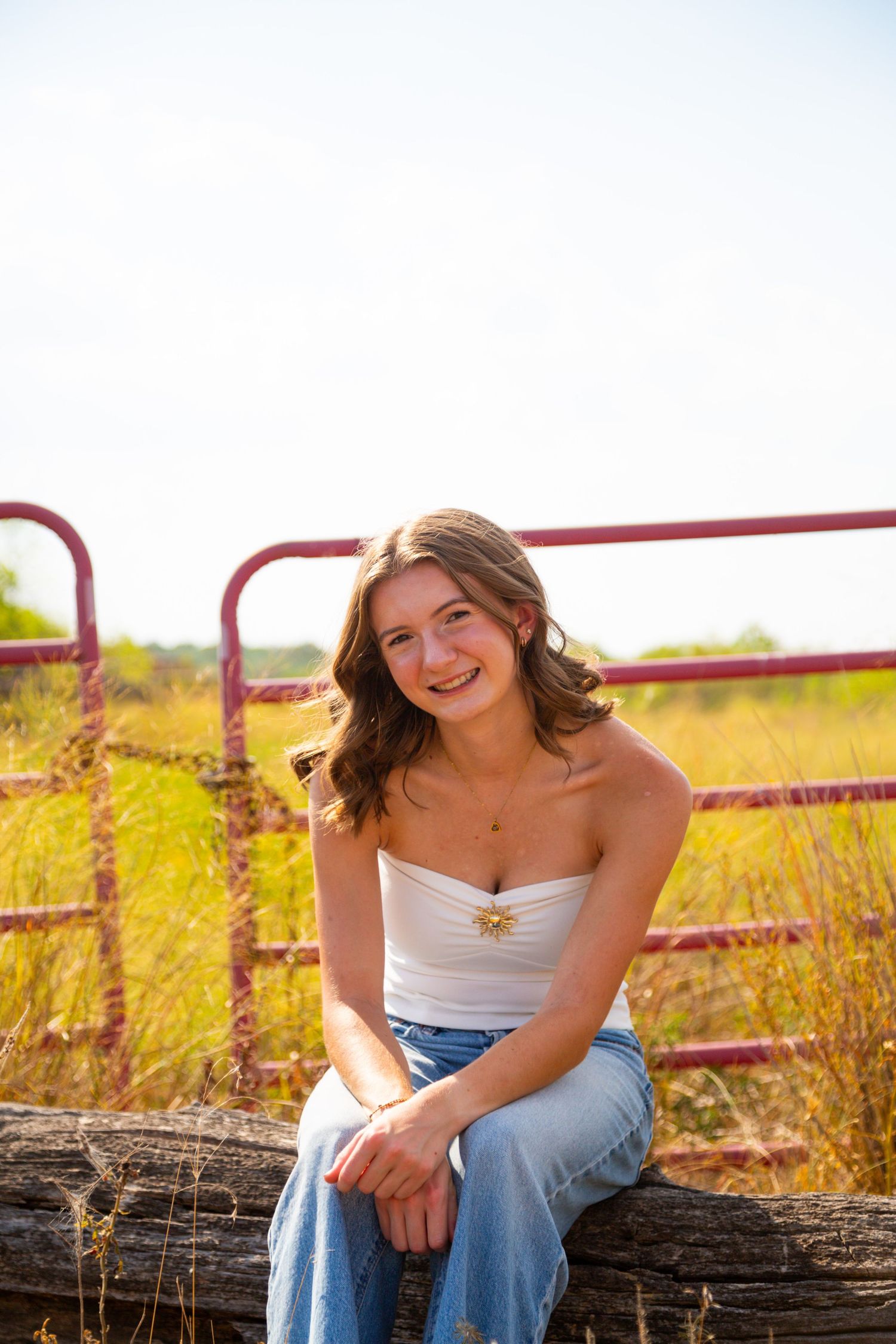 Smiling teenage girl in white tube top and jeans sits on a log fence near a rusty red gate in a sunny field.