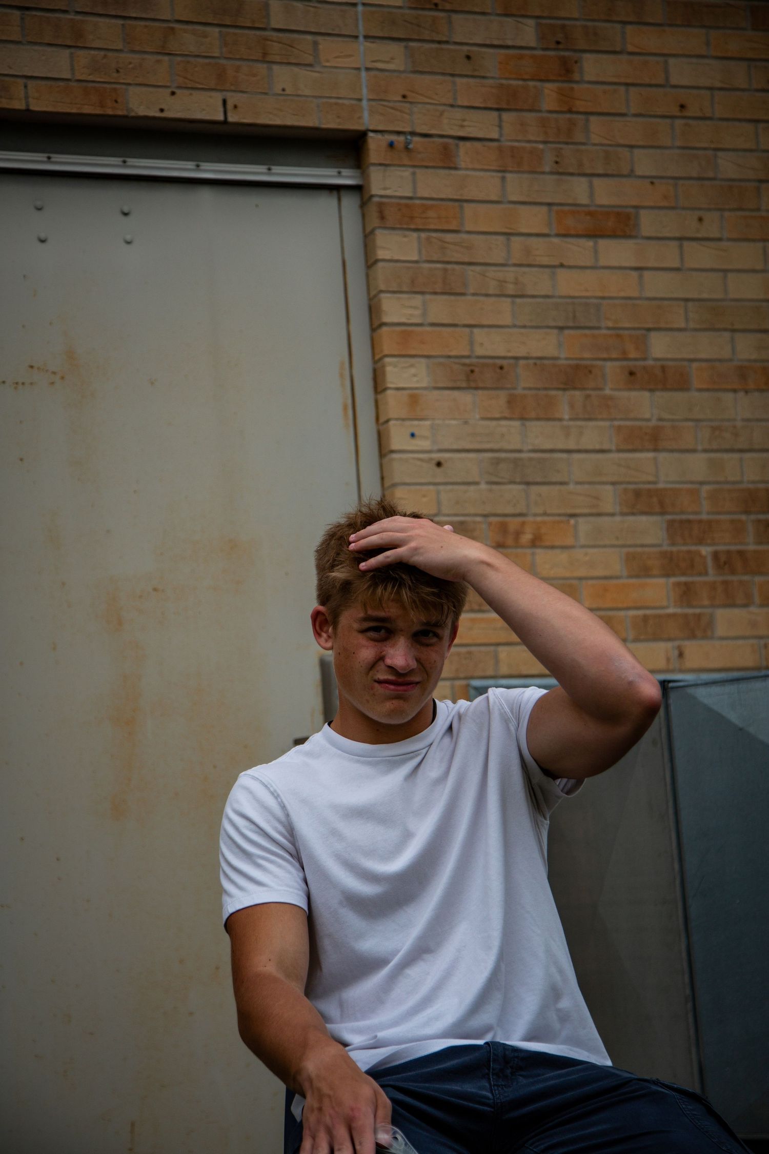 Young teenage boy in white t-shirt sits against brick wall touching his hair.