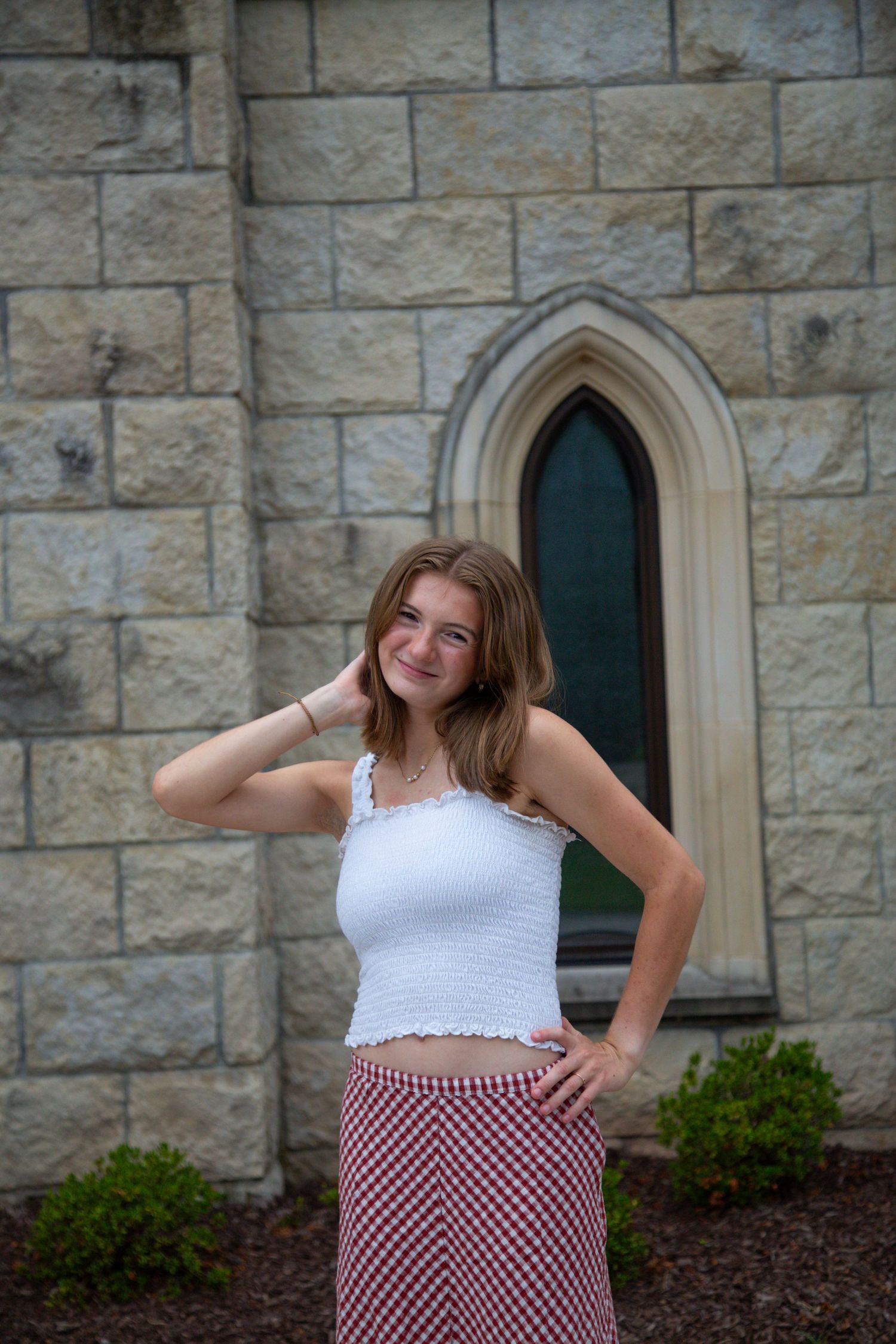Smiling teen girl in white crop top and red plaid skirt posing by Gothic stone church window.