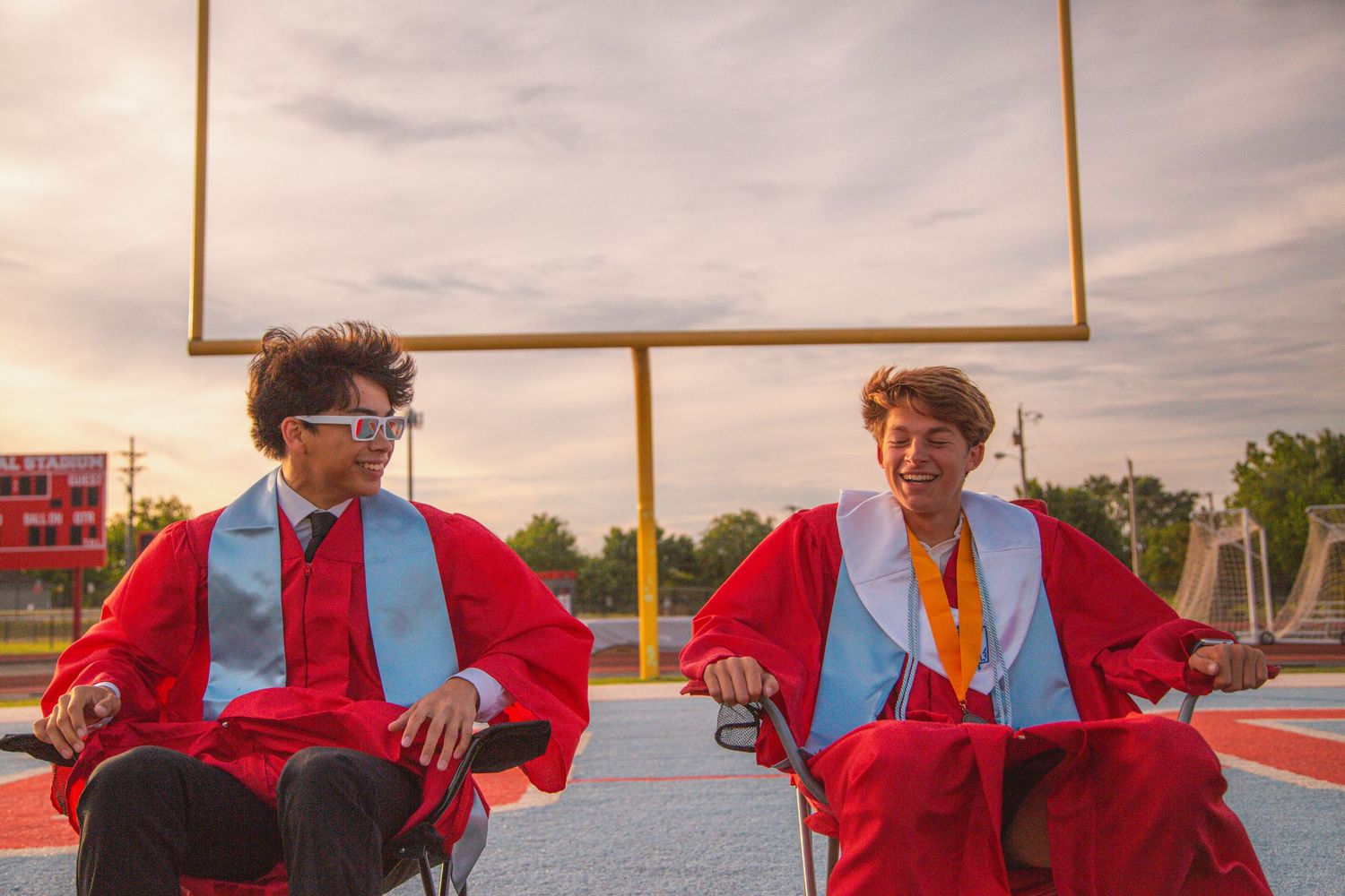 Two graduates in red gowns sit in chairs on football field at sunset wearing fun glasses.