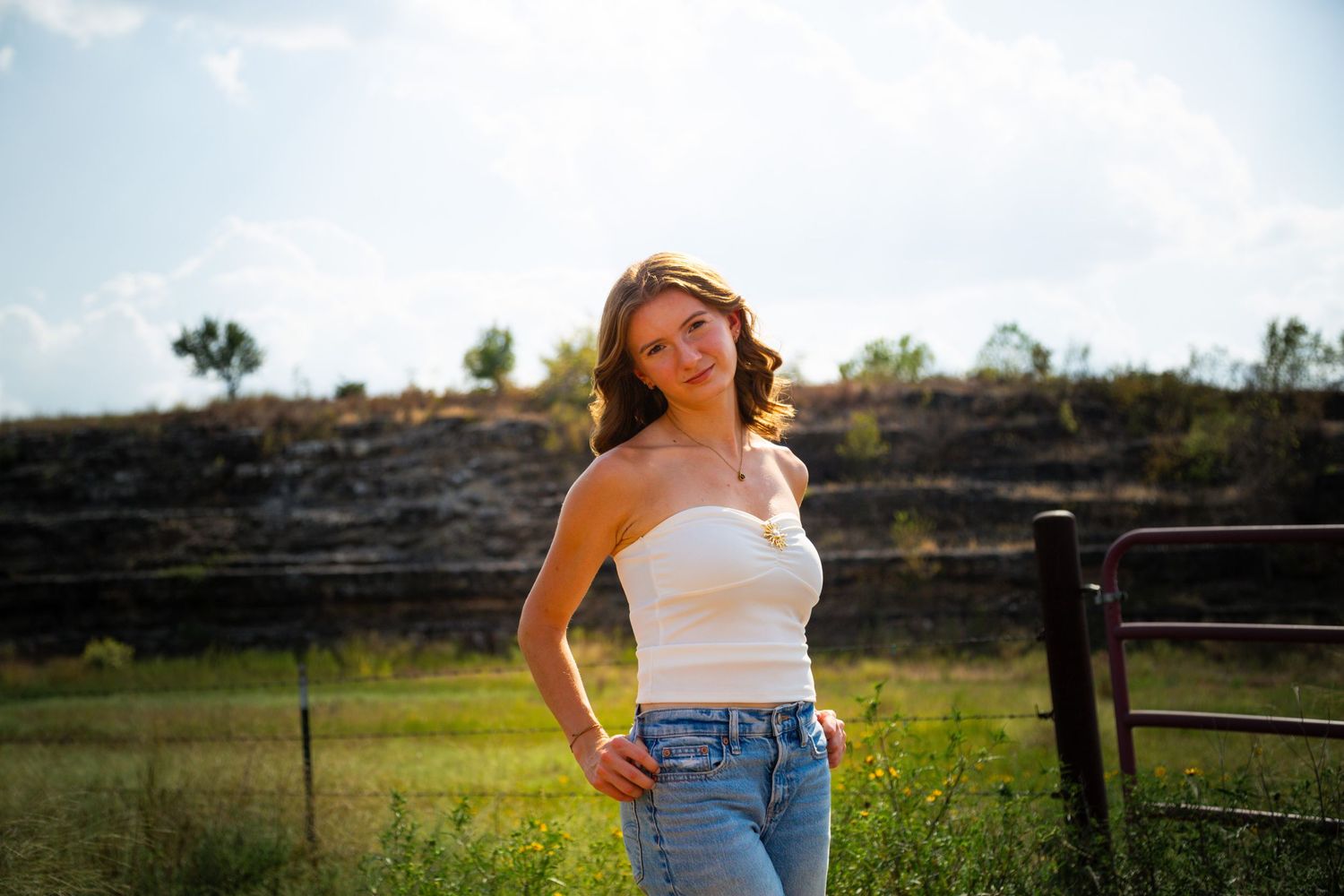 Young woman in white tube top and jeans posing outdoors in sunny green field near wooden fence.