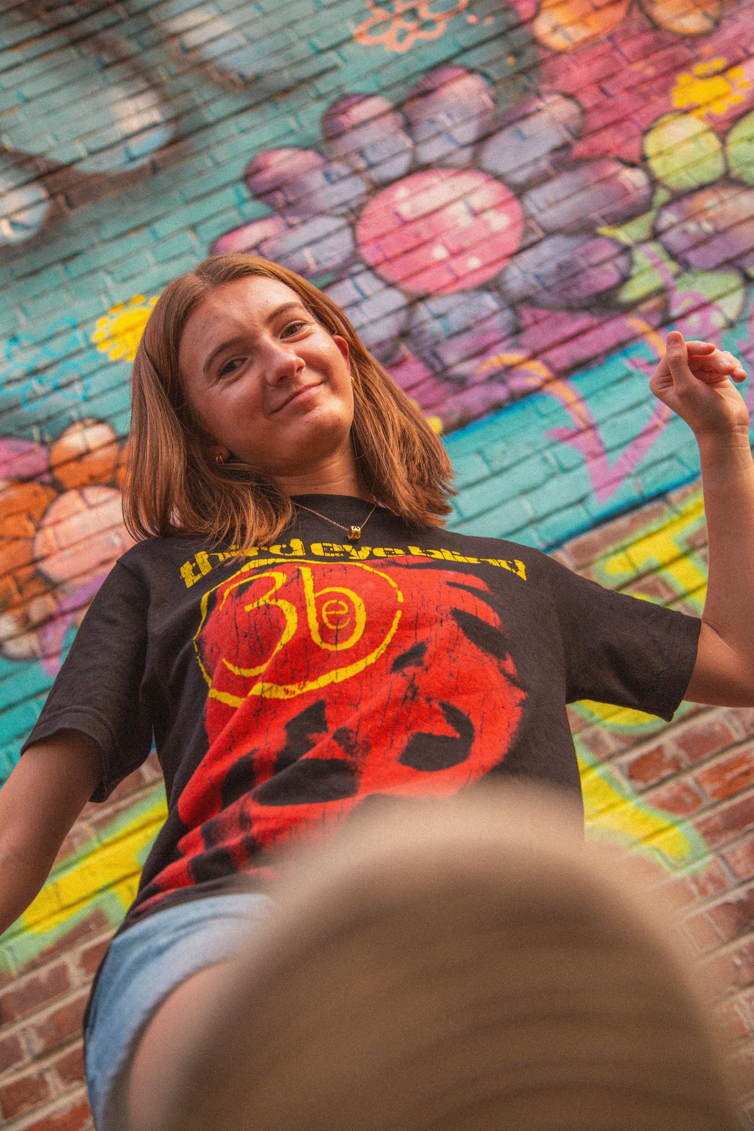 Young woman in a Third Eye Blind band tee smiles against a vibrant colorful graffiti mural backdrop.