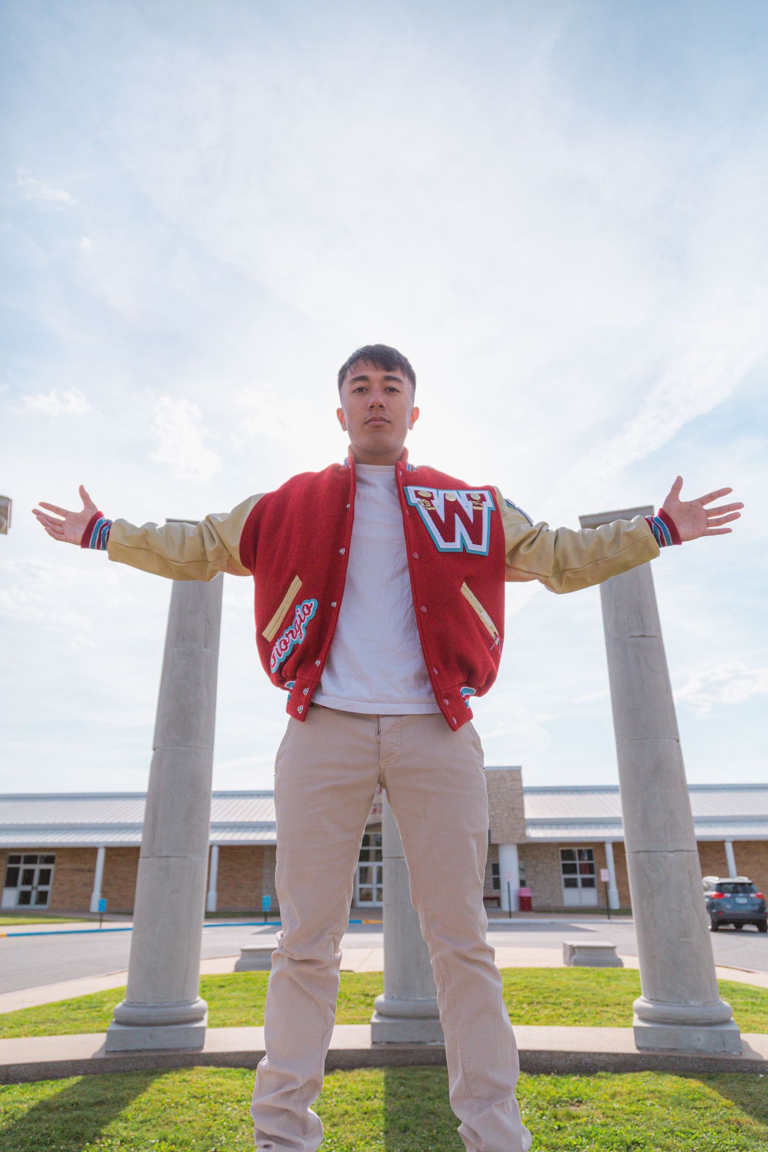A young man in a red varsity jacket spreads his arms wide standing between columns at Webb City High School.