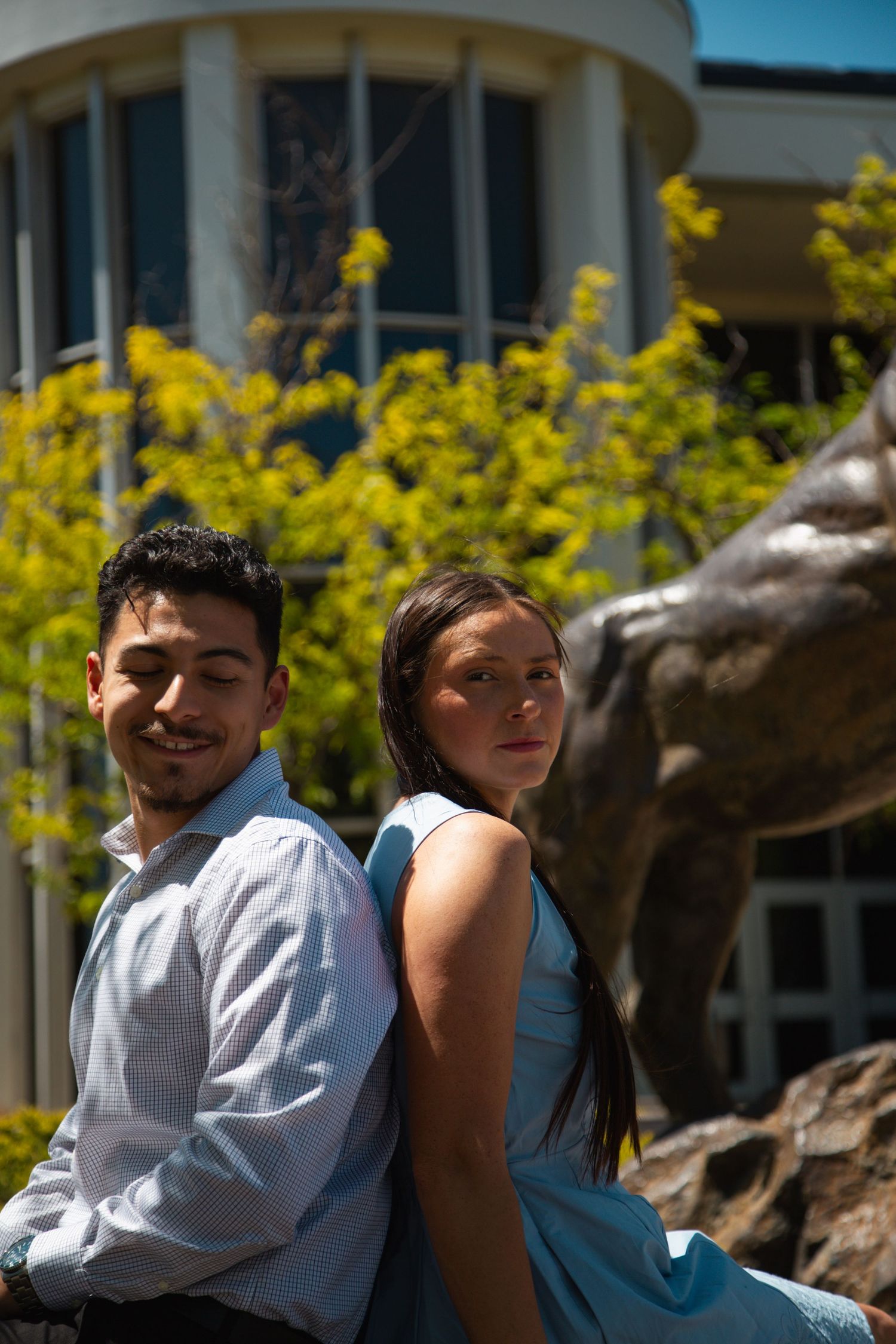 Young couple posing back-to-back outdoors near a bronze statue and building.