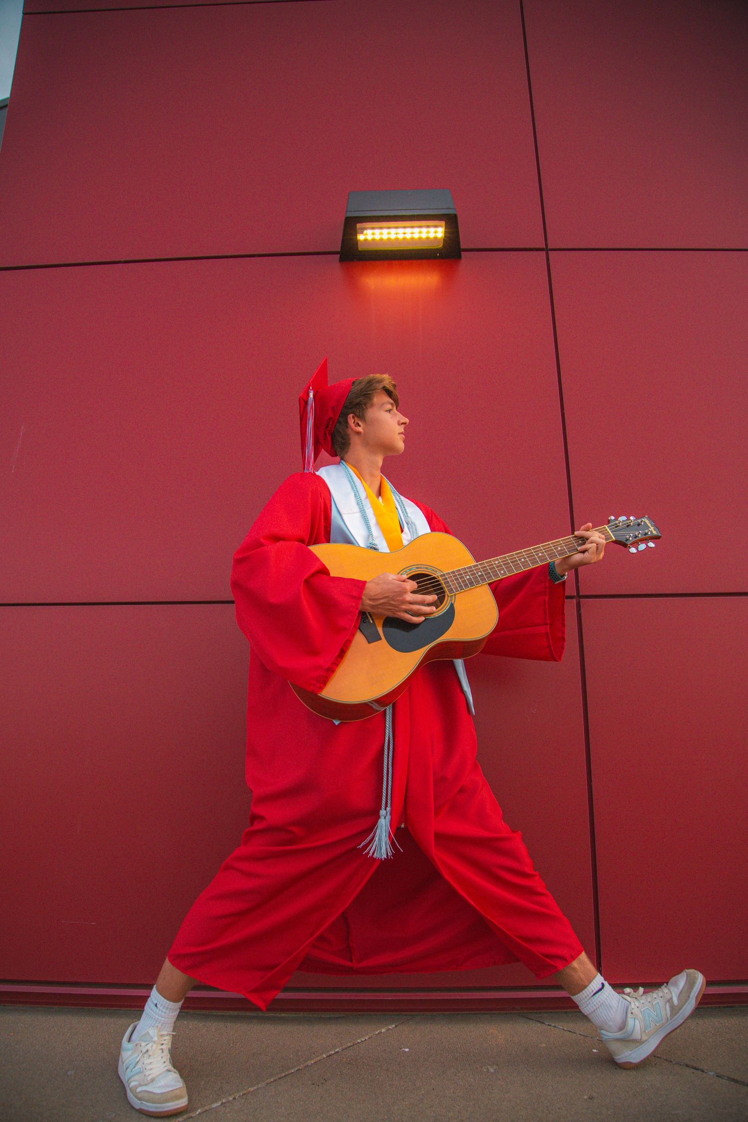 Graduate in red cap and gown playing acoustic guitar against a red wall outdoors.