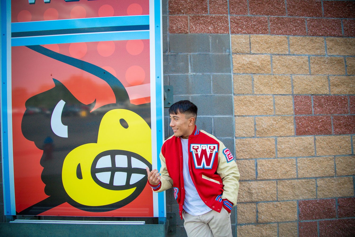 Young man in varsity jacket leaning against colorful school mascot mural.