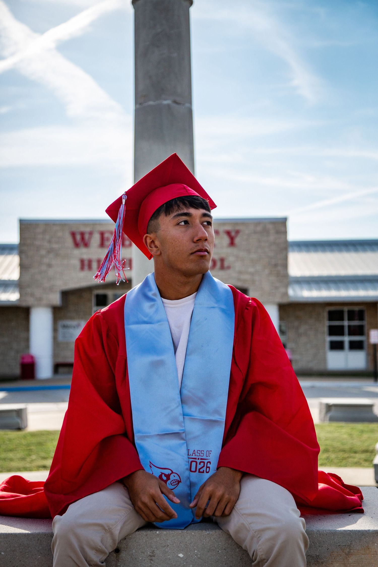 Graduate in red cap and gown sitting outside Webb City High School.
