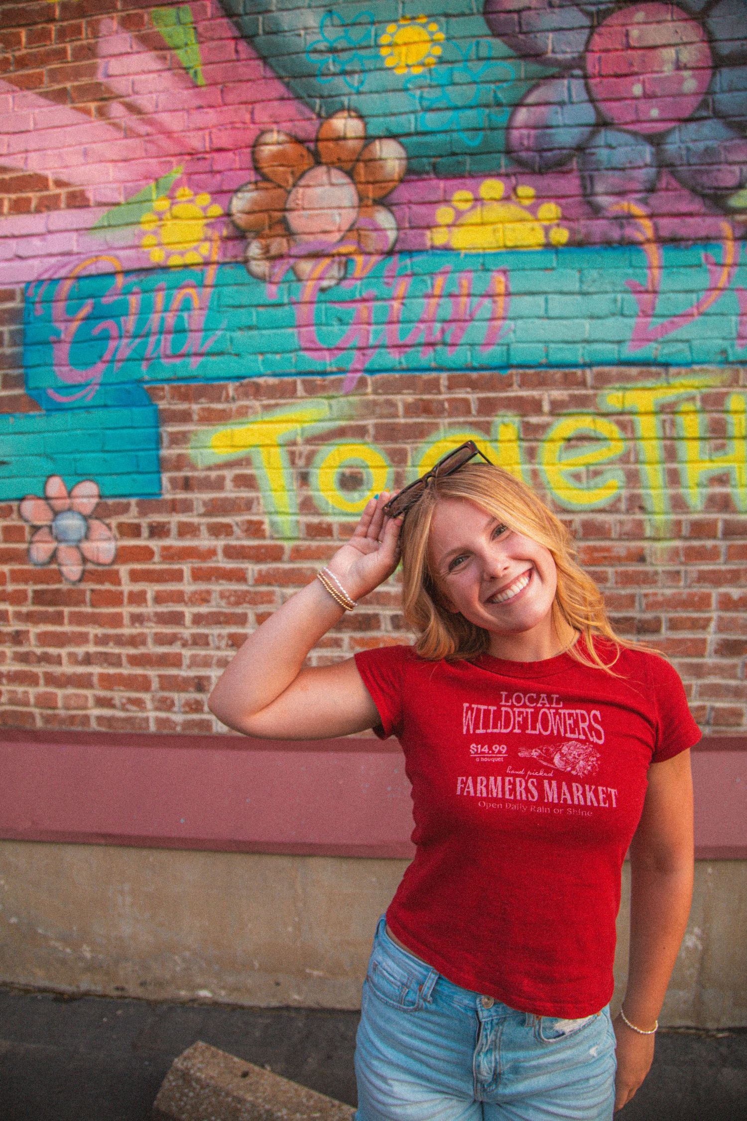 Cheerful blonde teen in red Wildflowers tee smiles before vibrant floral Together brick wall mural.