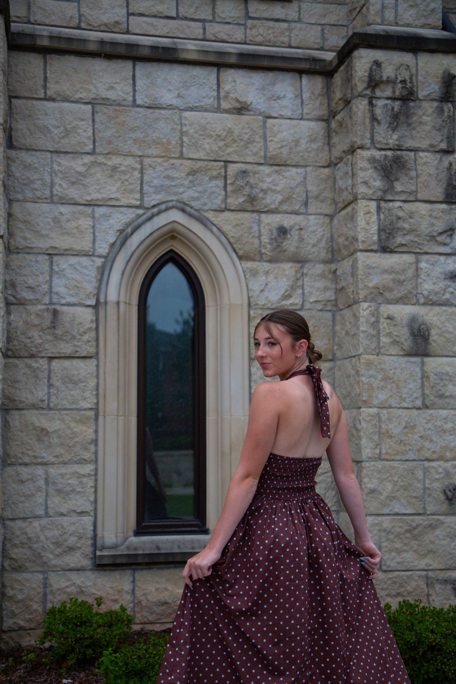 Young woman in burgundy polka dot halter dress poses beside Gothic stone church window.