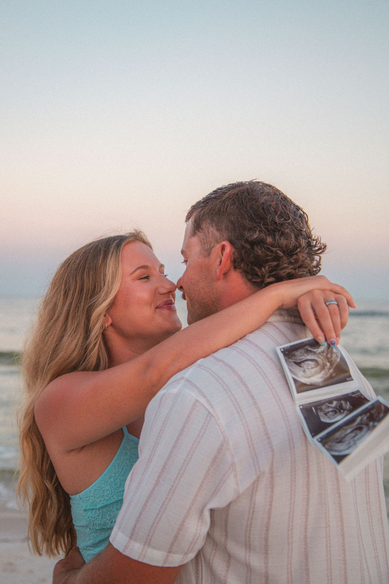 Couple embracing on beach at sunset holding ultrasound photos announcing pregnancy.