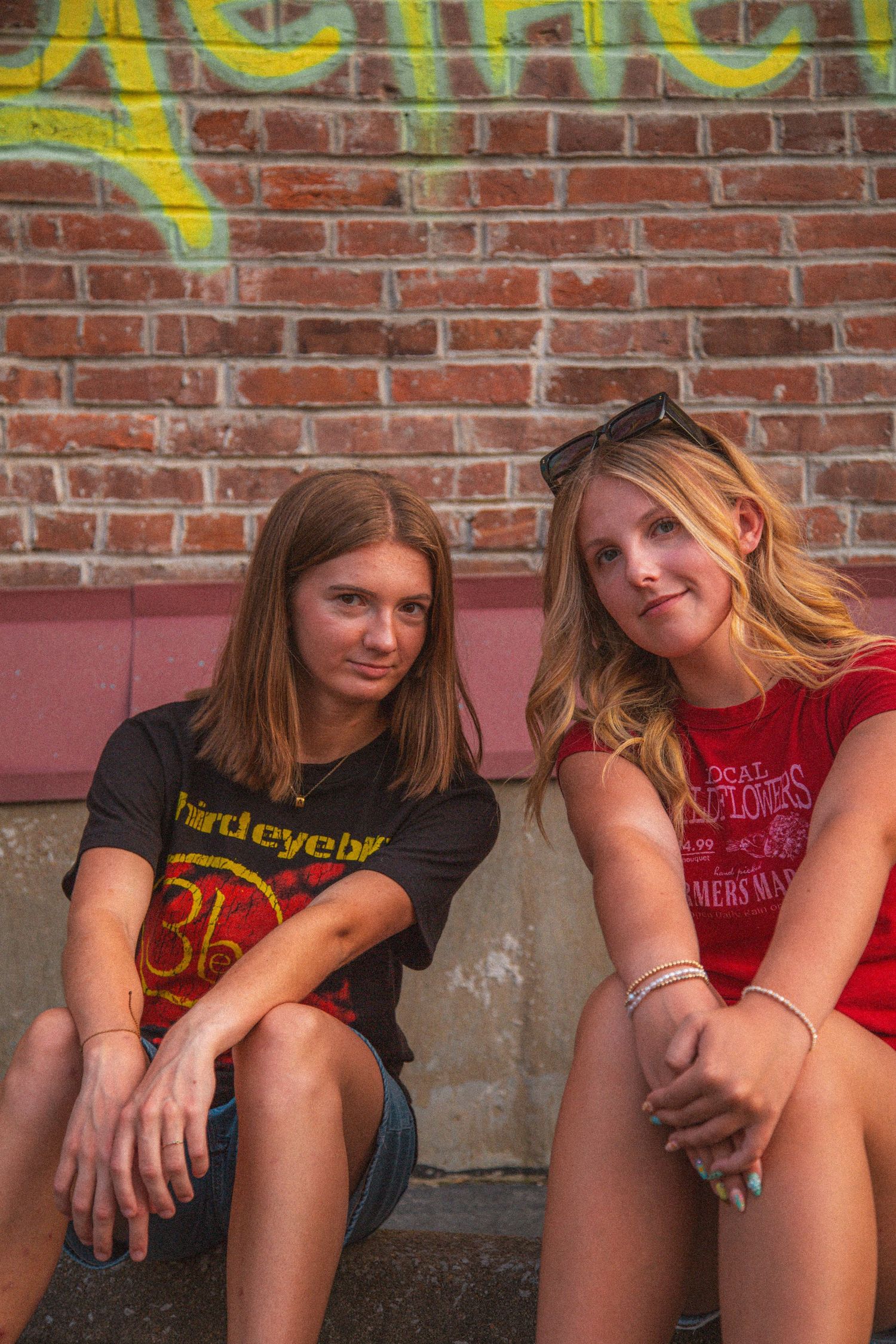 Two teenage girls in casual tees sit together against brick wall with colorful graffiti mural.
