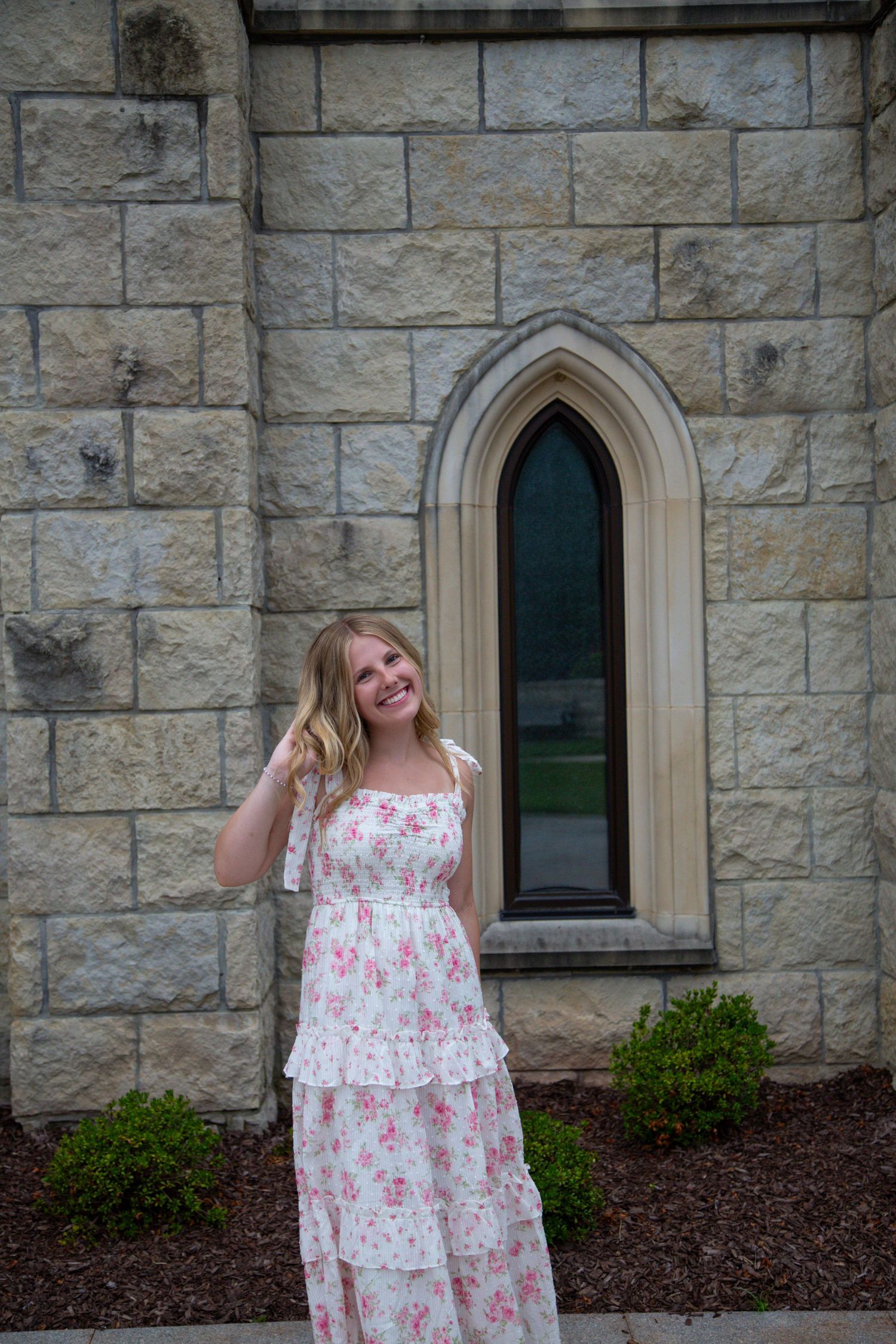 Smiling young woman in white floral maxi dress posing in front of stone church with Gothic arch window.
