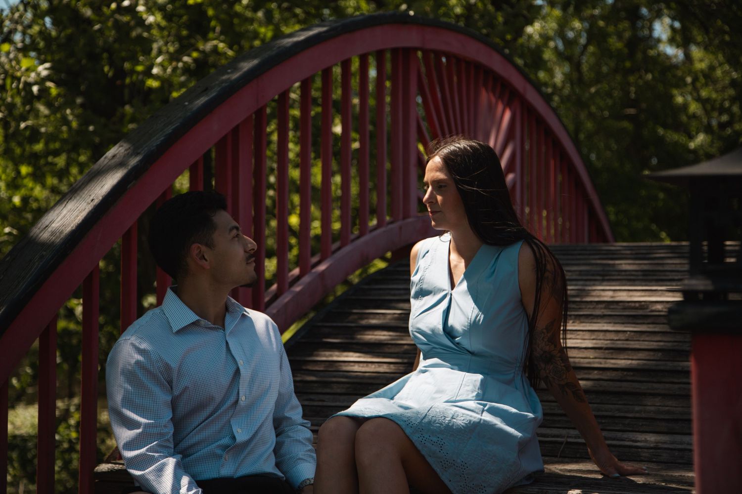 Young couple in blue outfits sharing an intimate conversation while sitting on red arched bridge.