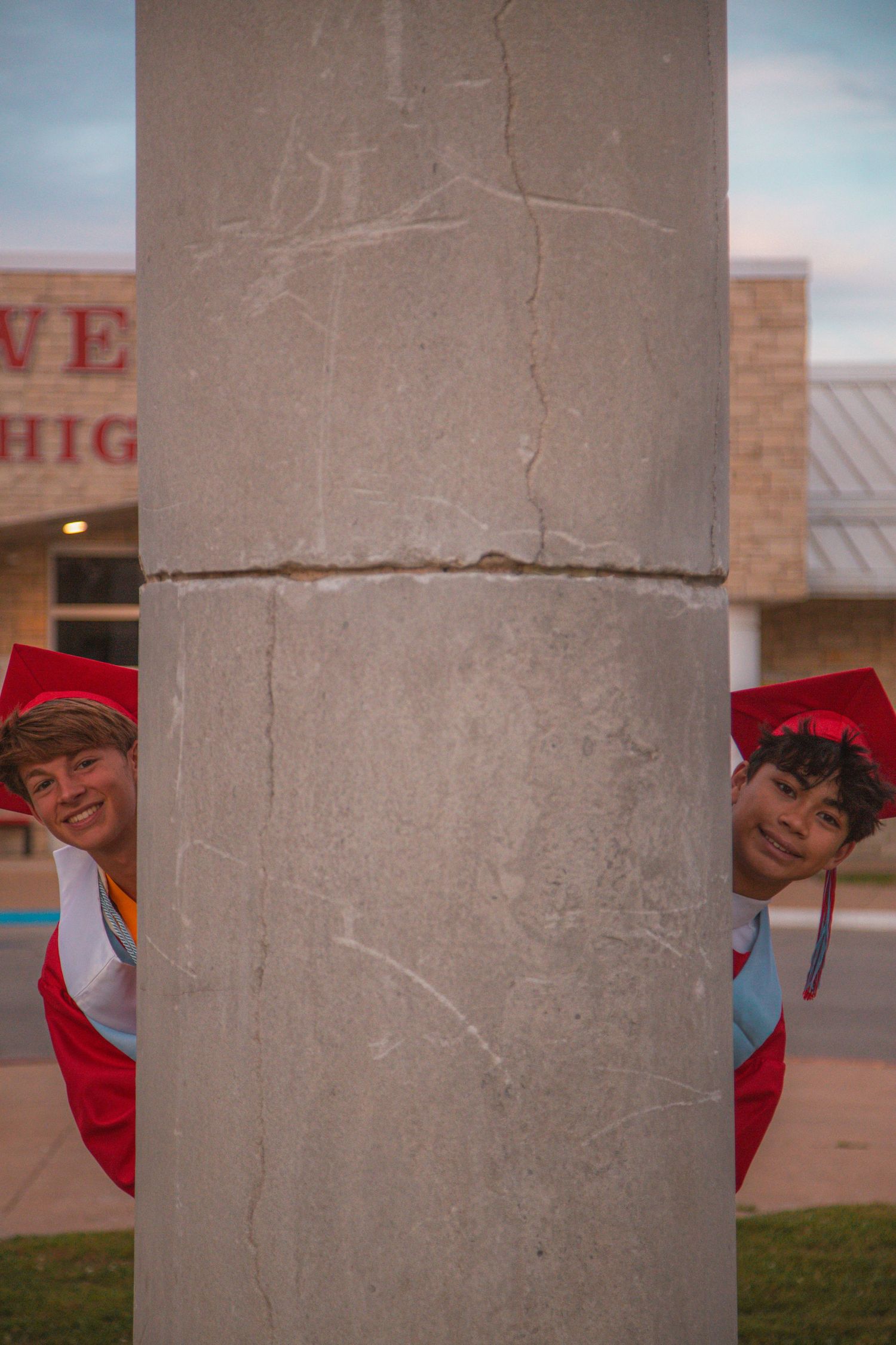 Two graduates in red caps and gowns playfully peeking out from behind a concrete pillar at Webb City High School.