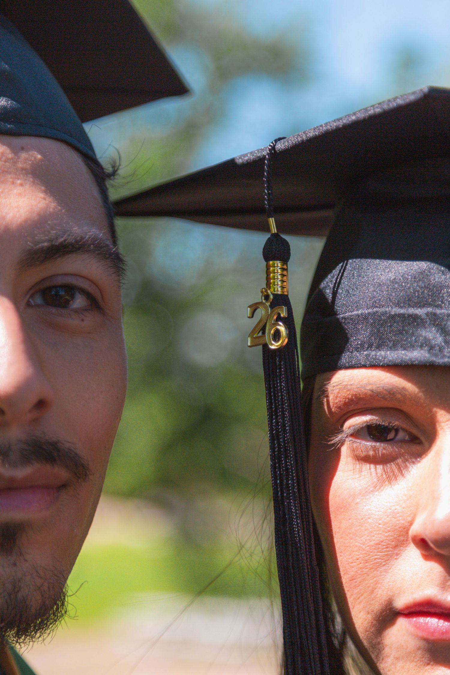 Close-up portrait of two graduates wearing black caps with gold 26 tassel charm in outdoor setting.
