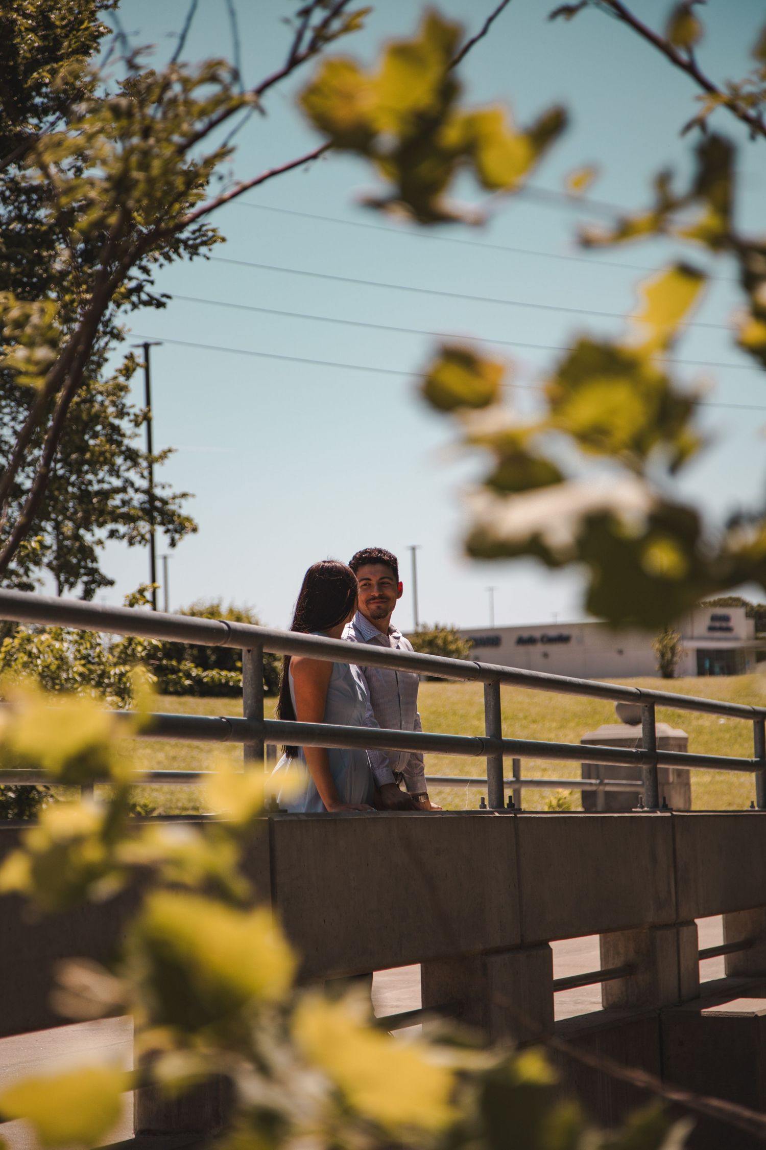 Couple standing close together on a bridge framed by green leaves against a clear blue sky.