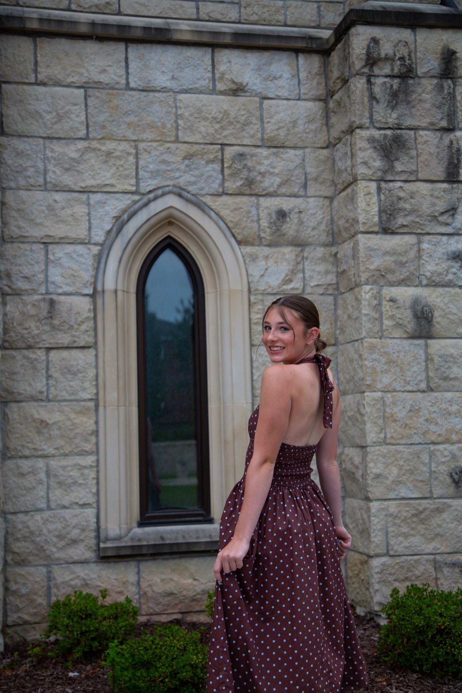 Smiling young woman in brown polka dot halter dress twirling near Gothic stone church window.
