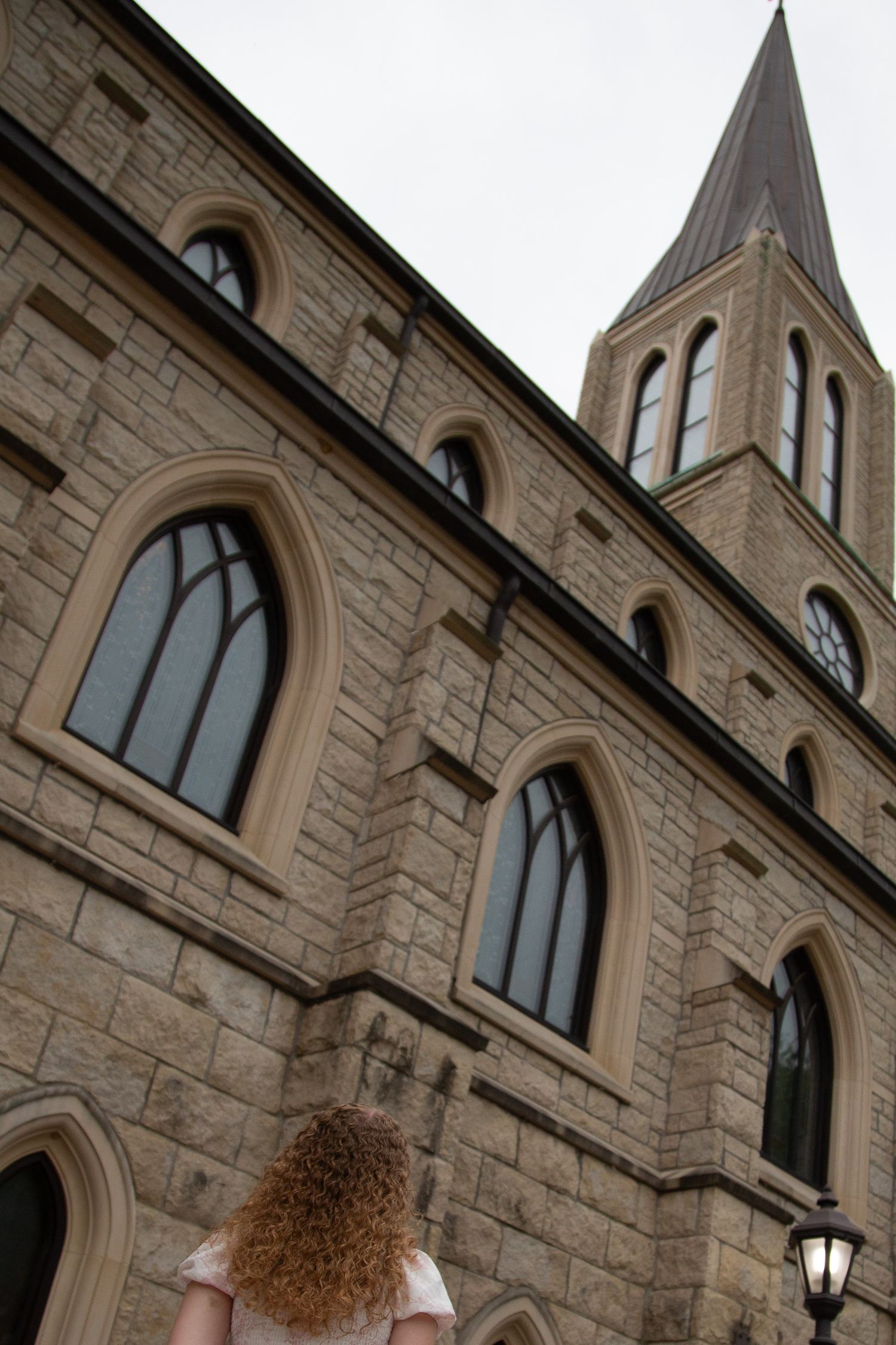 Curly red-haired girl looking up at tall gothic stone church with steeple.