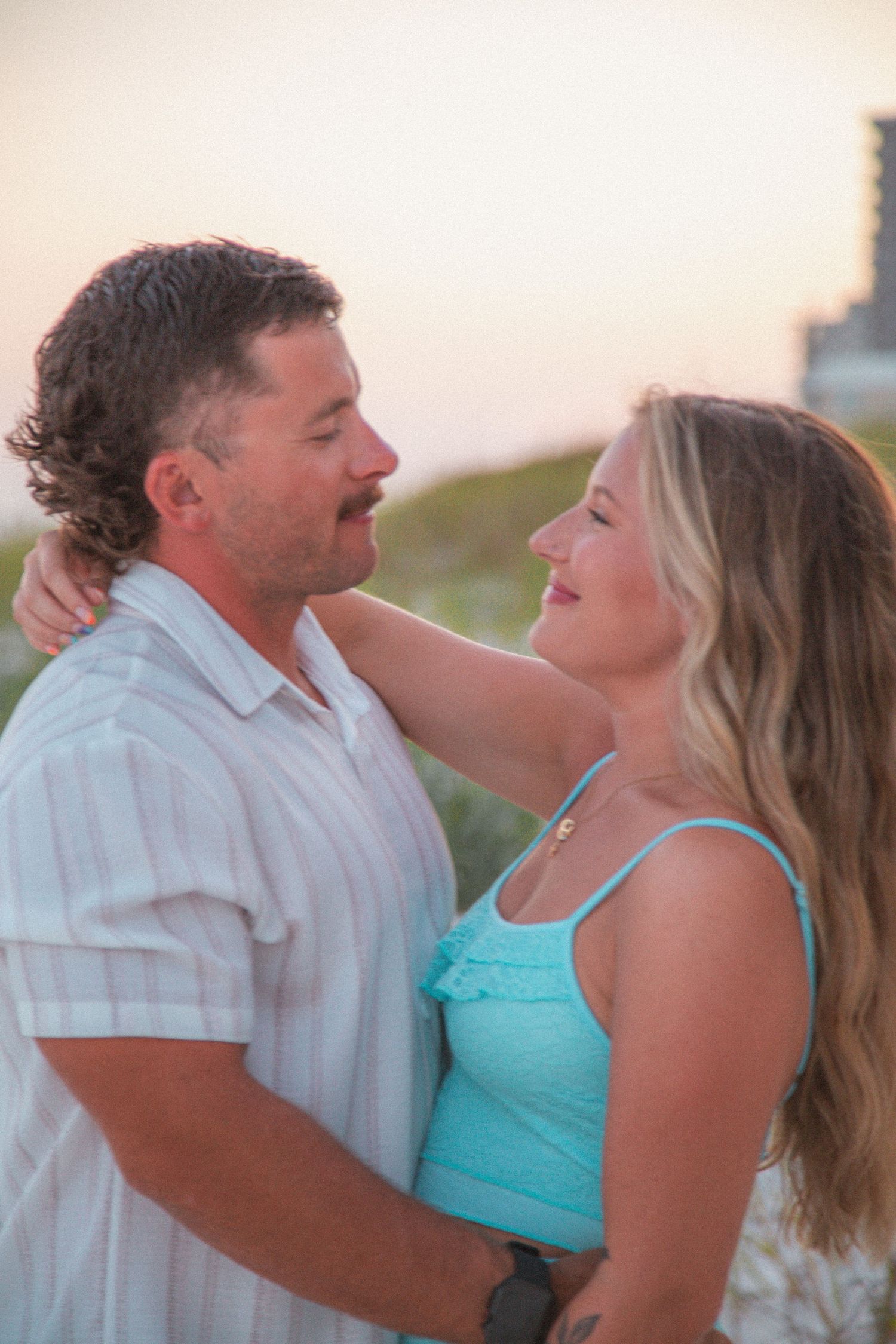 Couple sharing a romantic moment at sunset on the beach, woman in turquoise dress smiling up at man.