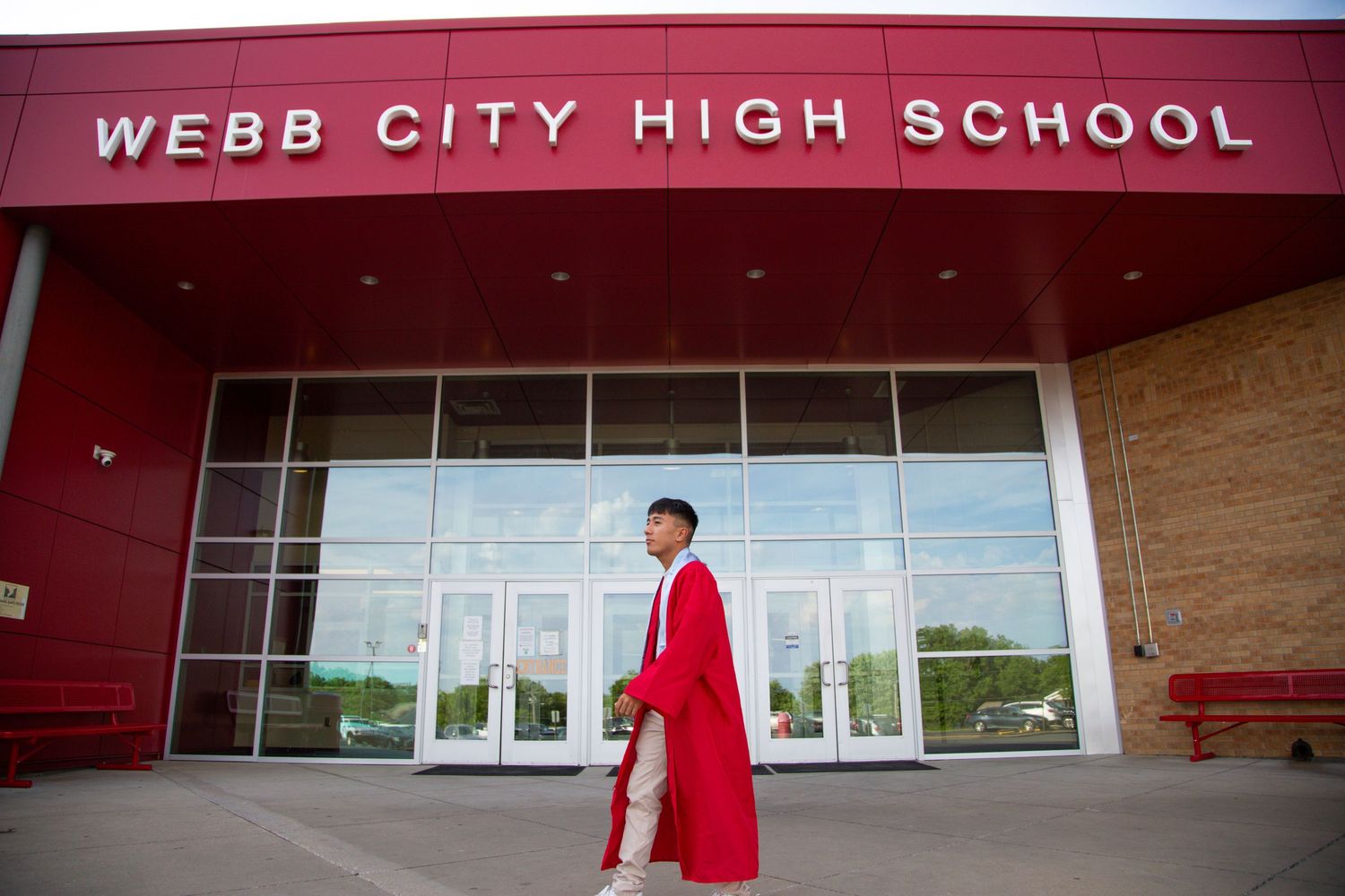 Graduate in red cap and gown walking in front of Webb City High School entrance on sunny day.