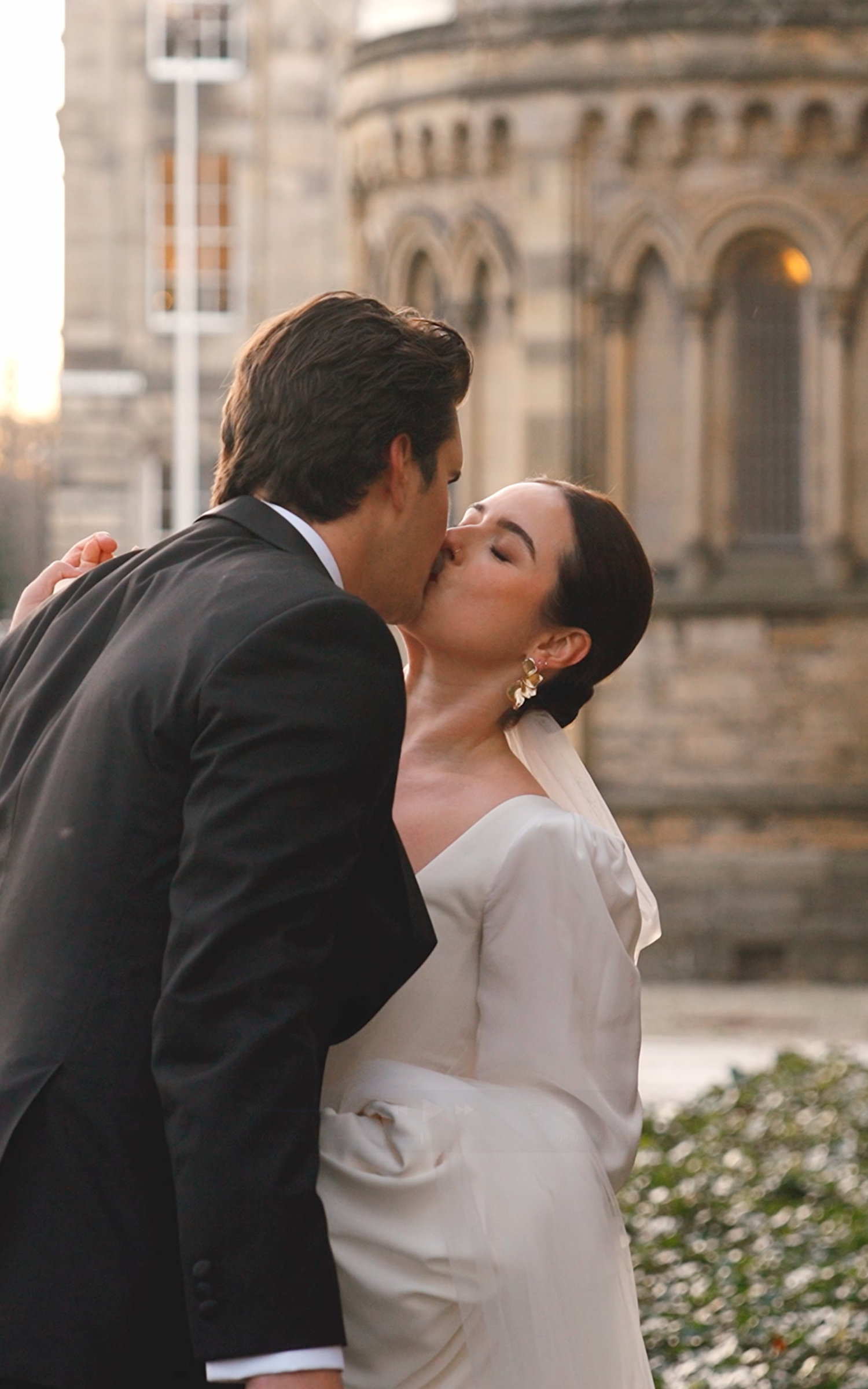 A newlywed couple shares a romantic kiss outside a historic stone church at golden hour.