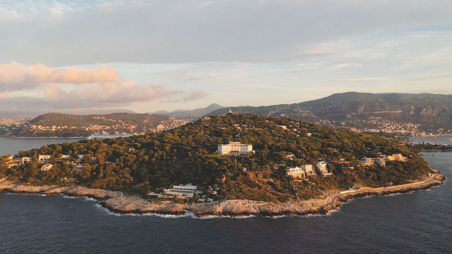 Aerial view of a green wooded peninsula with villas along a rocky Mediterranean coastline at sunset, with hazy mountains in the background.