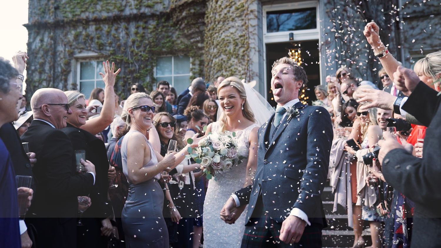 Bride and groom celebrate with confetti outside a stone venue surrounded by cheering guests.