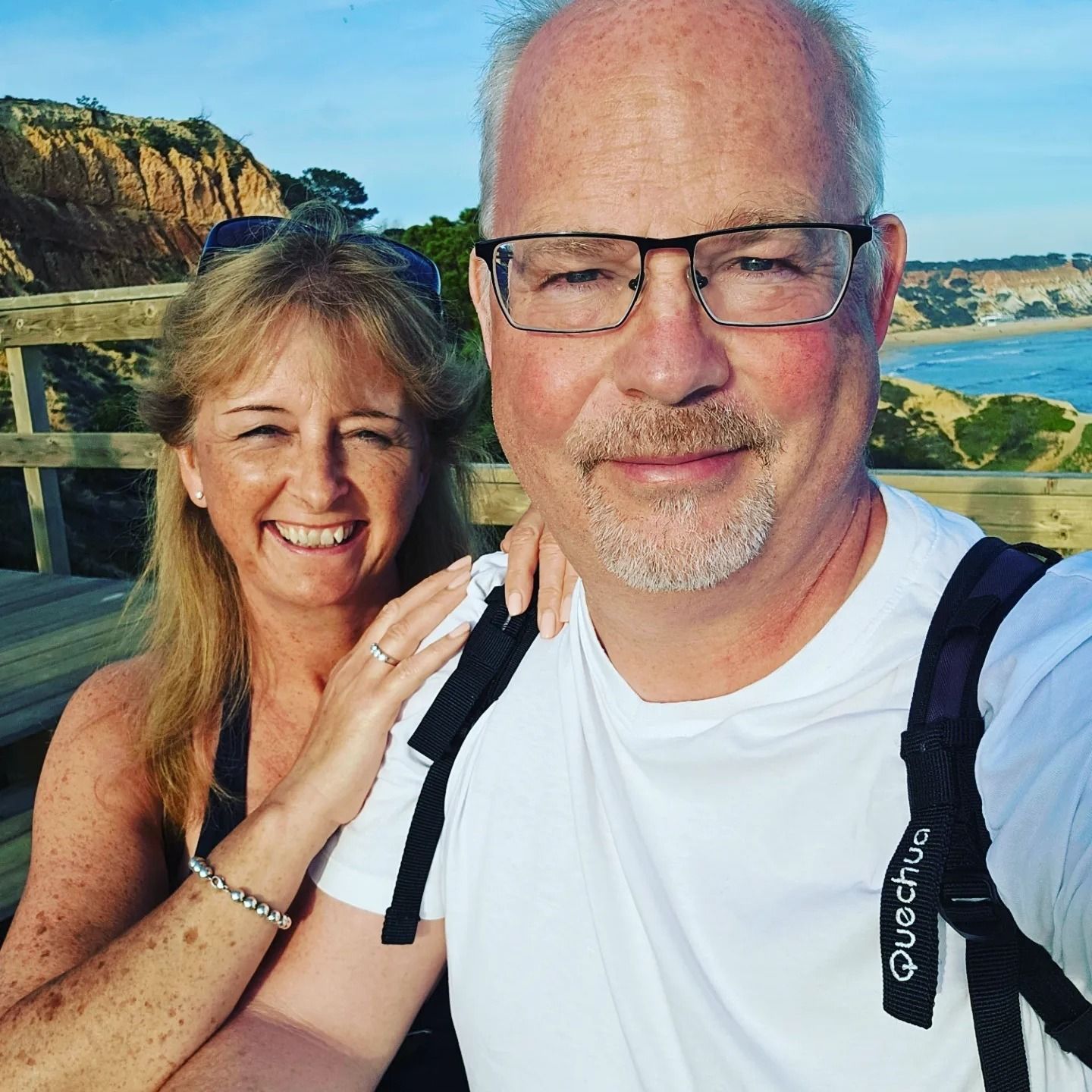 A smiling middle-aged couple takes a selfie on a wooden bridge with coastal cliffs and ocean in the background.