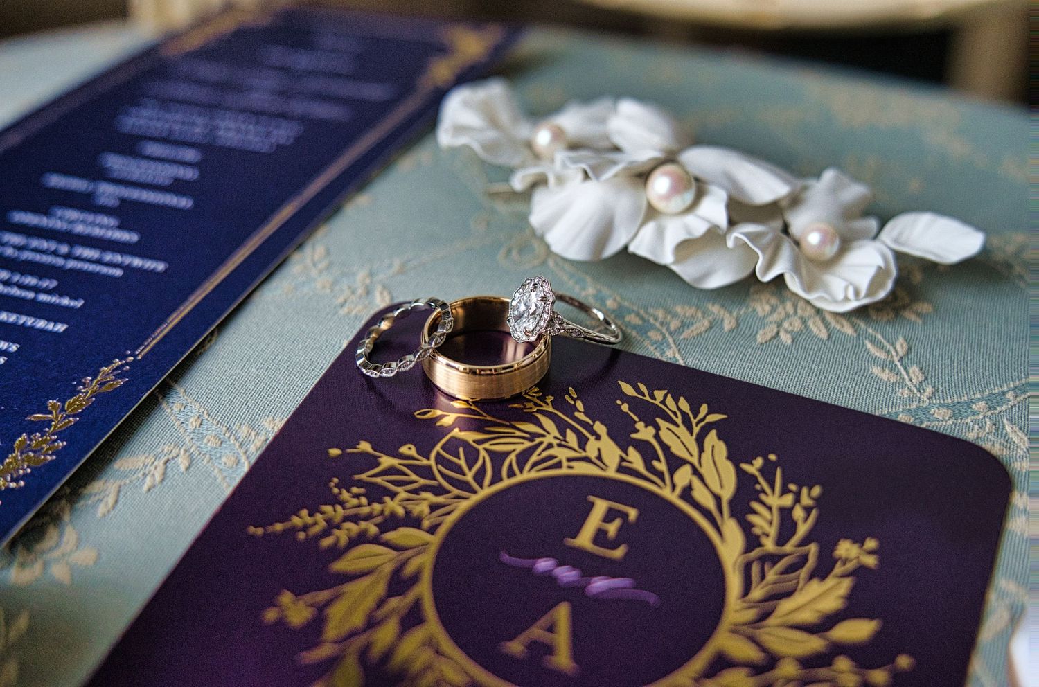 Elegant wedding stationery with gold monogram rings and floral details on a teal surface.