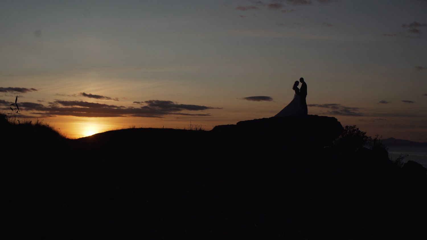 Silhouette of wedding couple standing on rocky hilltop during colorful sunset.