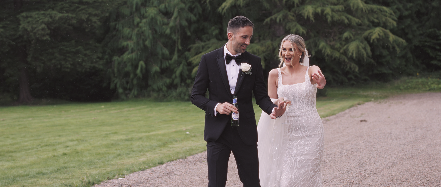 Elegant bride and groom walking hand in hand along a tree-lined path, holding champagne.