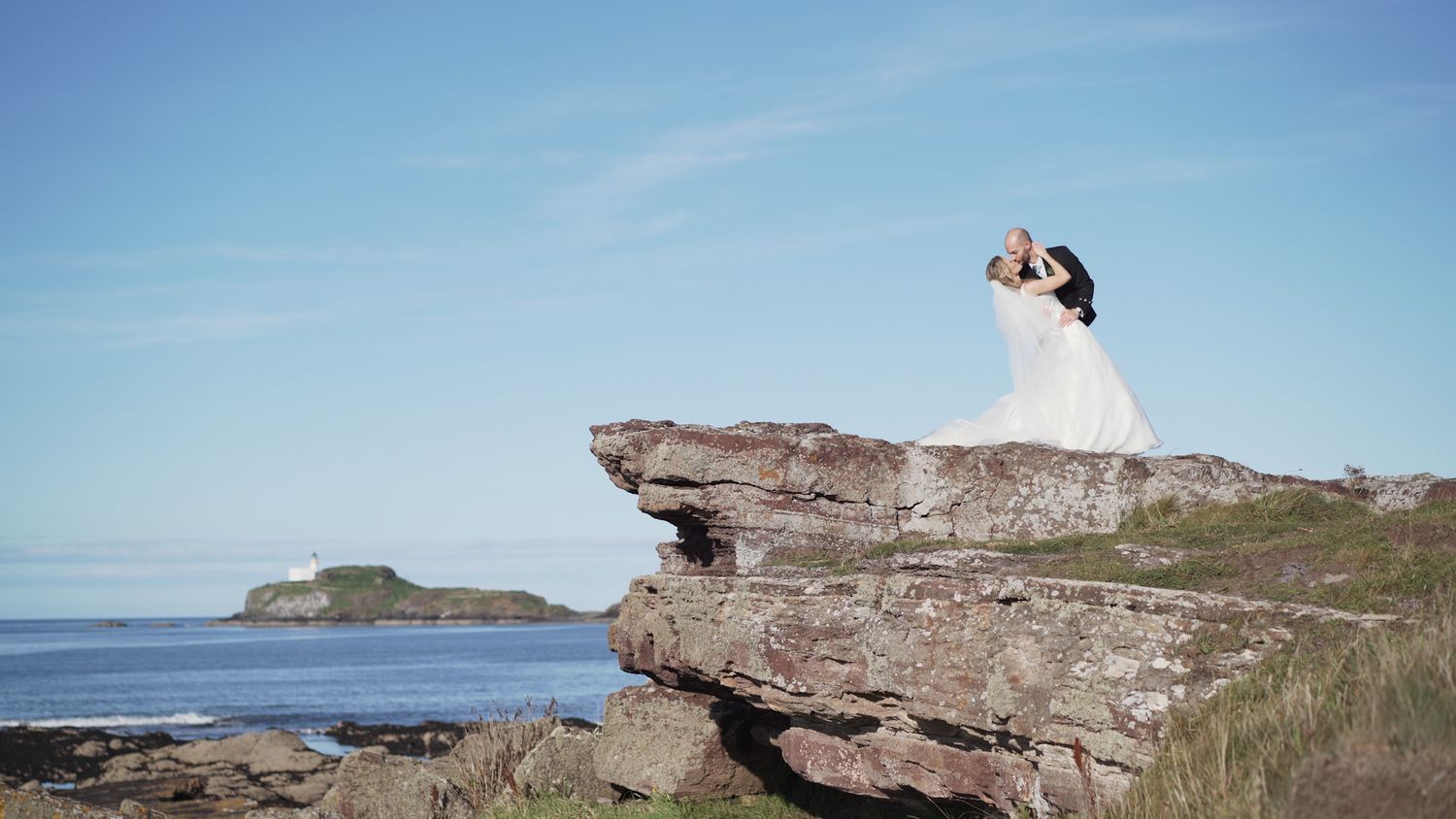 Wedding couple sharing a romantic dip kiss on coastal rocks with ocean backdrop.