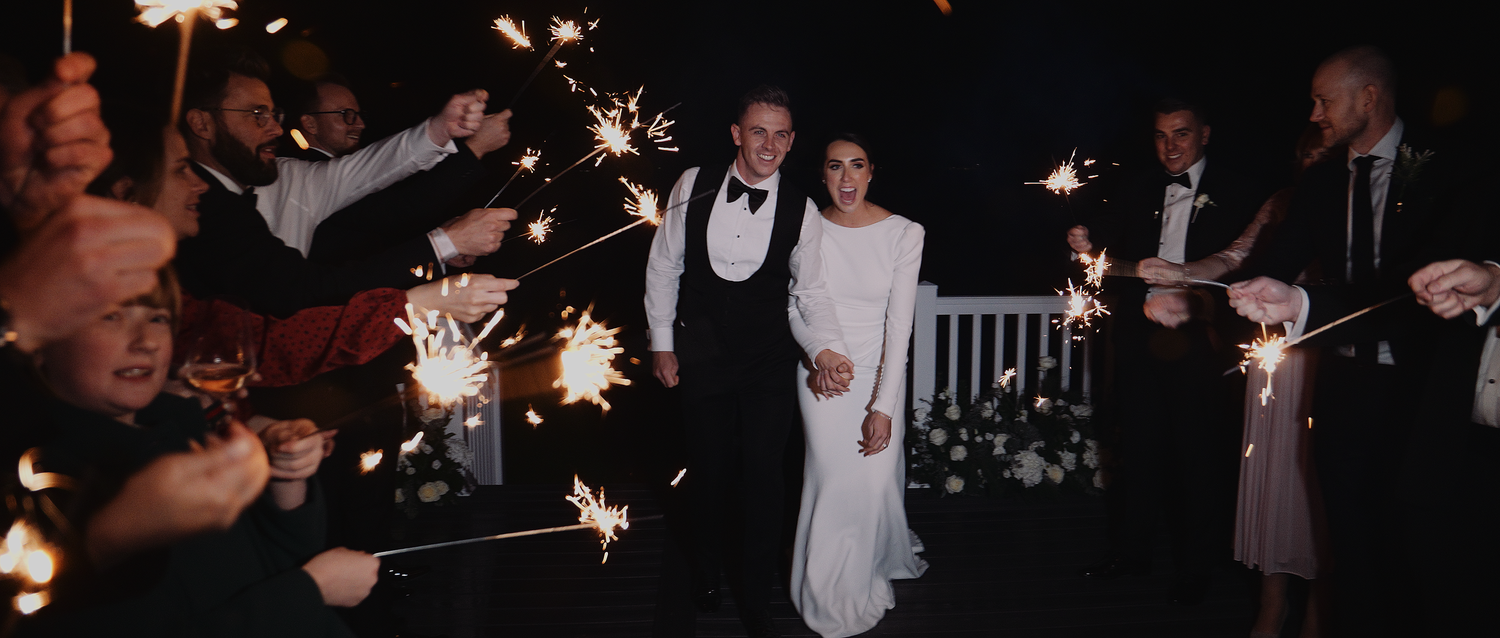 Wedding couple walking through sparkler send-off with guests at night celebration.