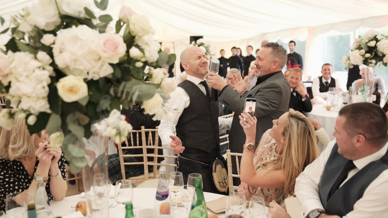Wedding guests applauding during heartfelt speech in elegant white floral decorated marquee.