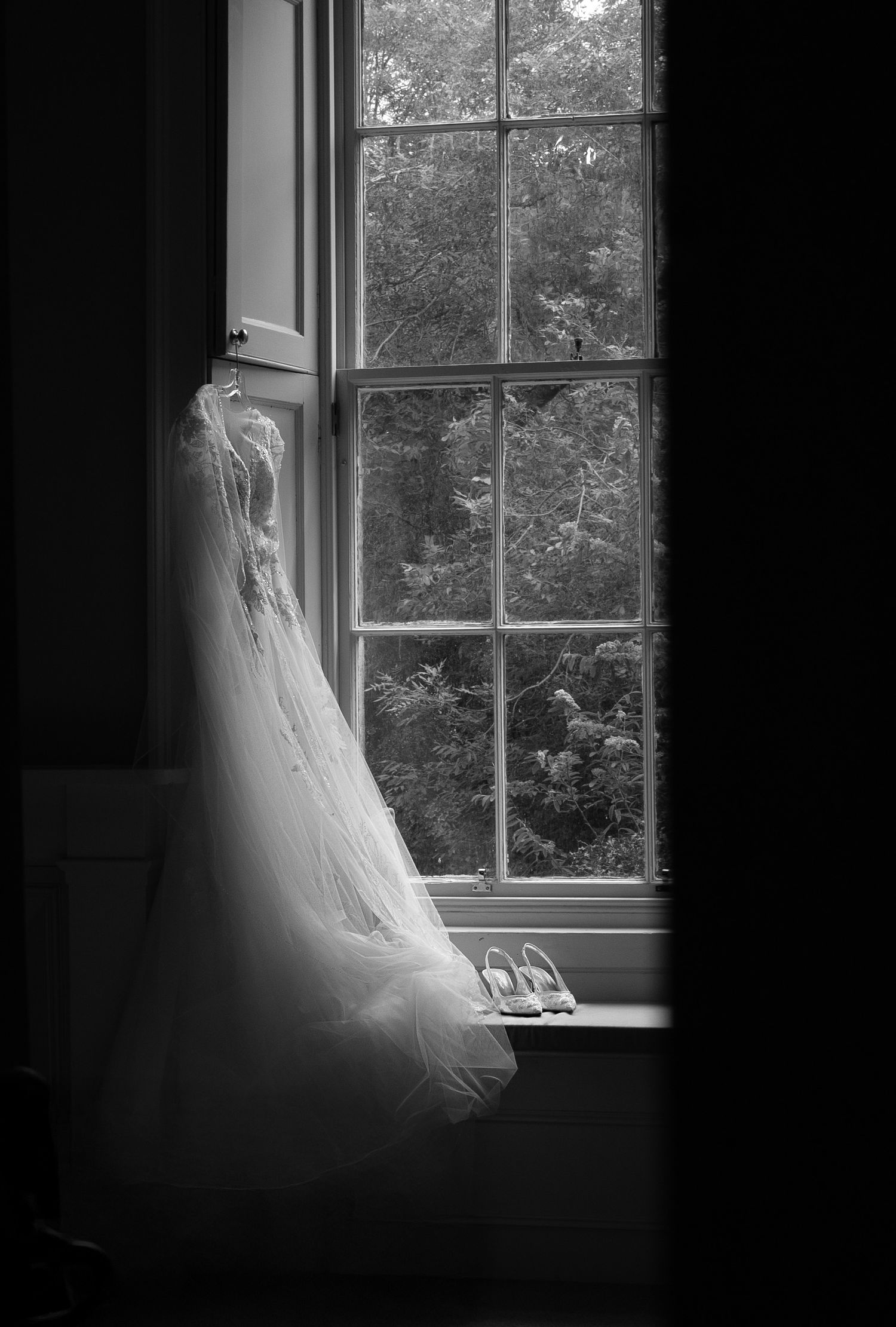 Black and white photo of a wedding dress and heels by a large window.