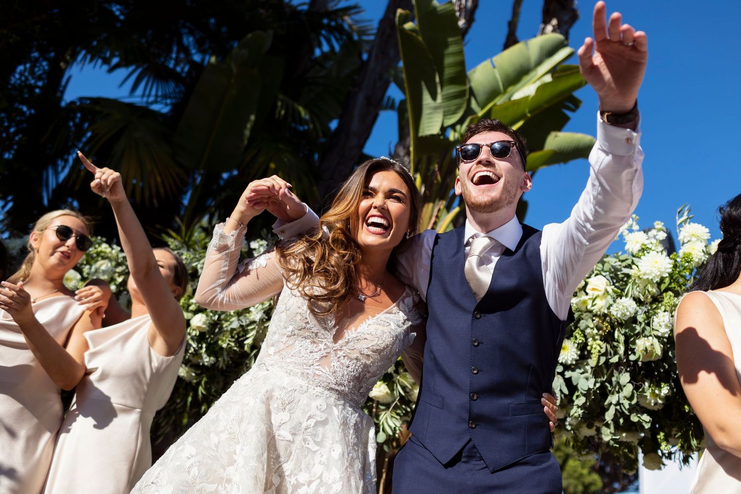 Joyful newlyweds celebrating outdoors with guests cheering holding white floral bouquets.