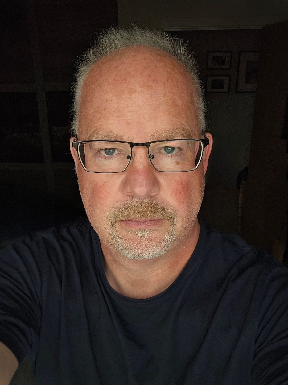 Close-up selfie of a middle-aged man with glasses and short gray hair indoors.