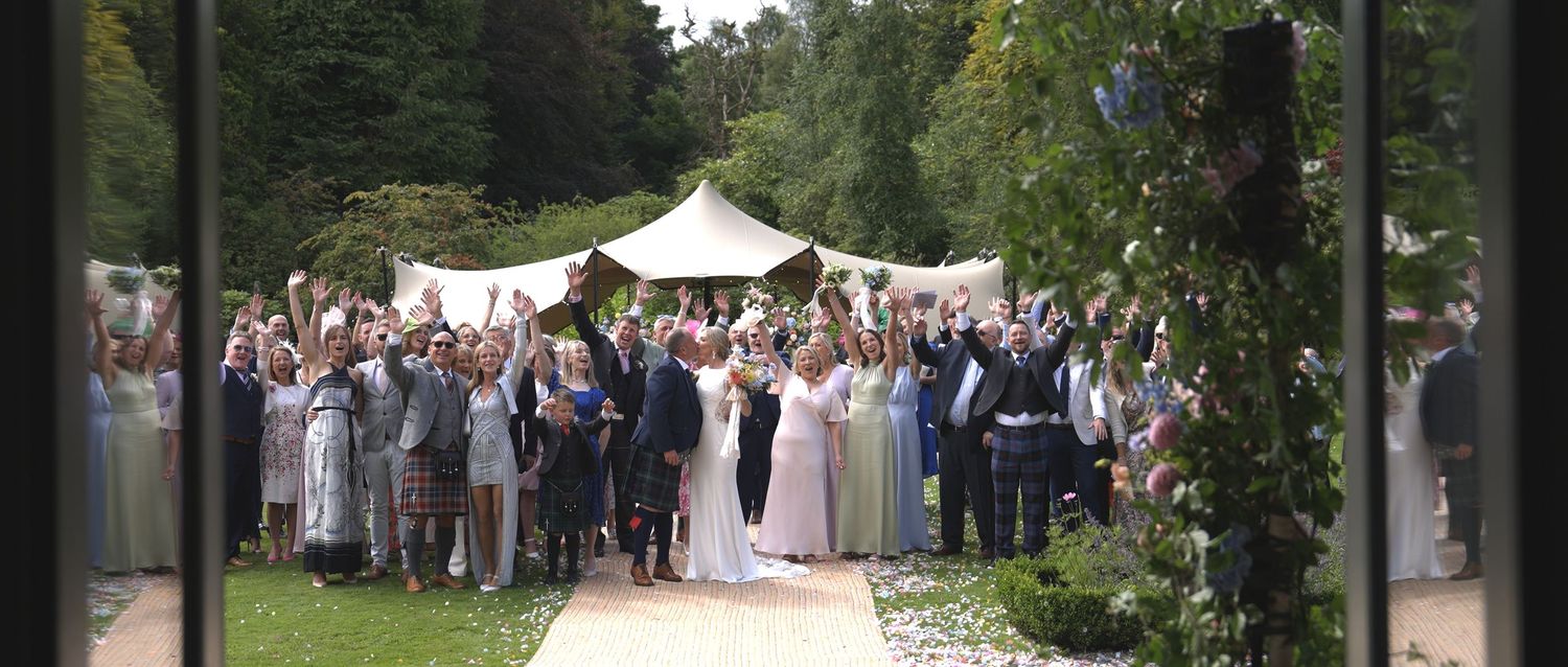 Large wedding party group photo outdoors under marquee tent surrounded by lush greenery.