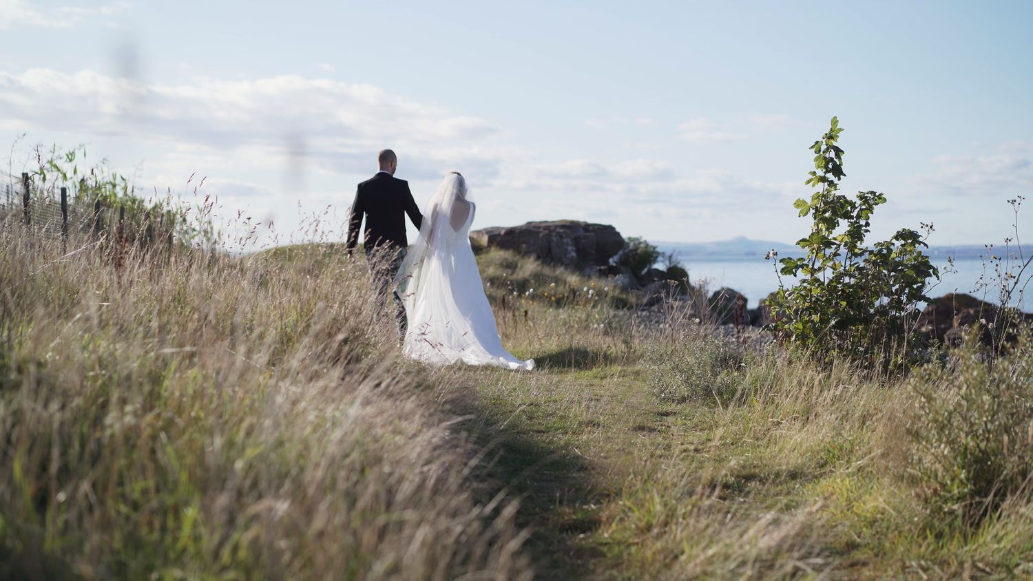 Wedding couple walking hand in hand through tall grass with ocean views behind.