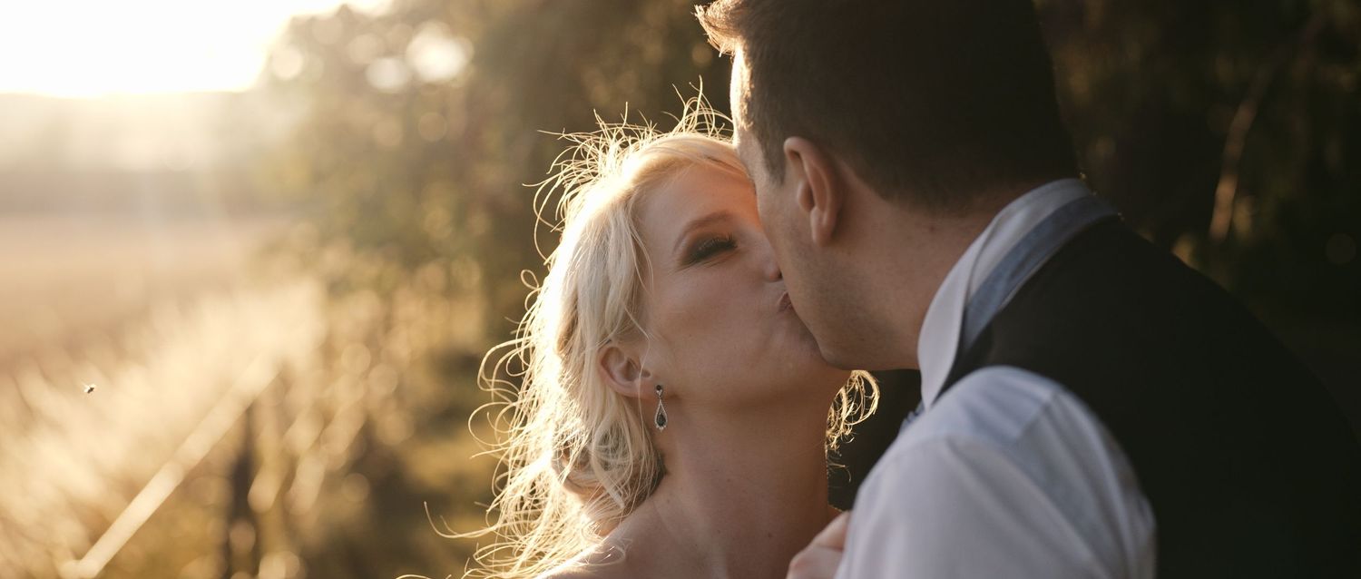 Bride and groom sharing a romantic kiss in warm golden sunset light in an outdoor setting.