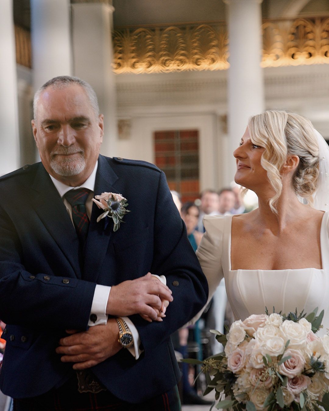 Bride walking down the aisle arm in arm with her father in a grand columned ceremony venue.