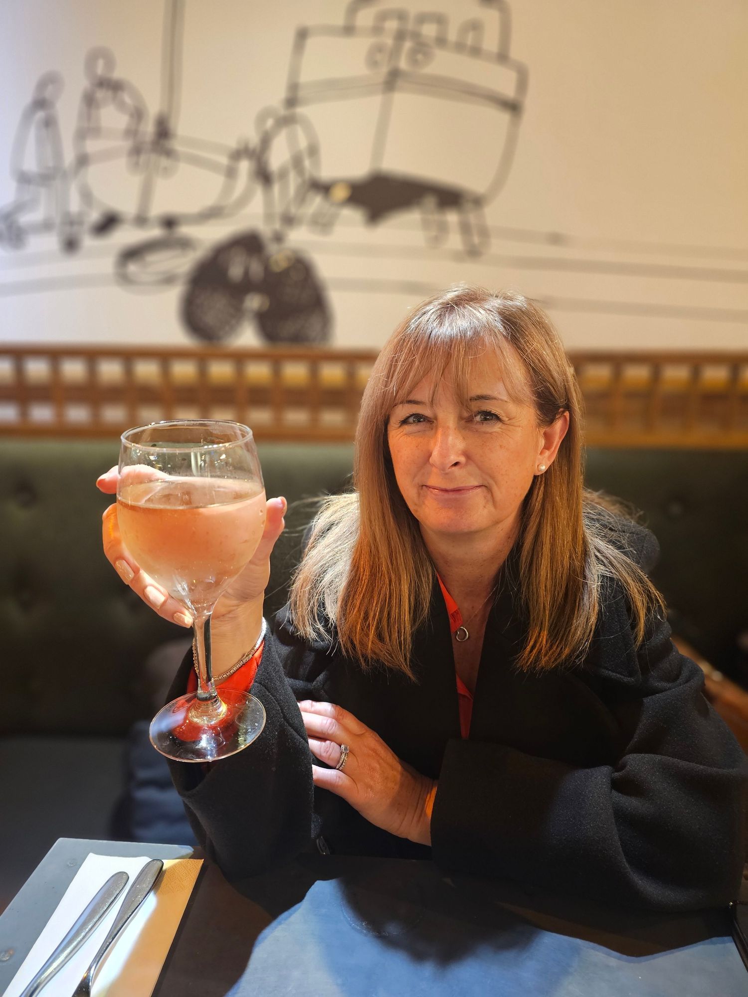 A woman raises a glass of rose wine in a cozy restaurant booth with illustrated wall art behind her.