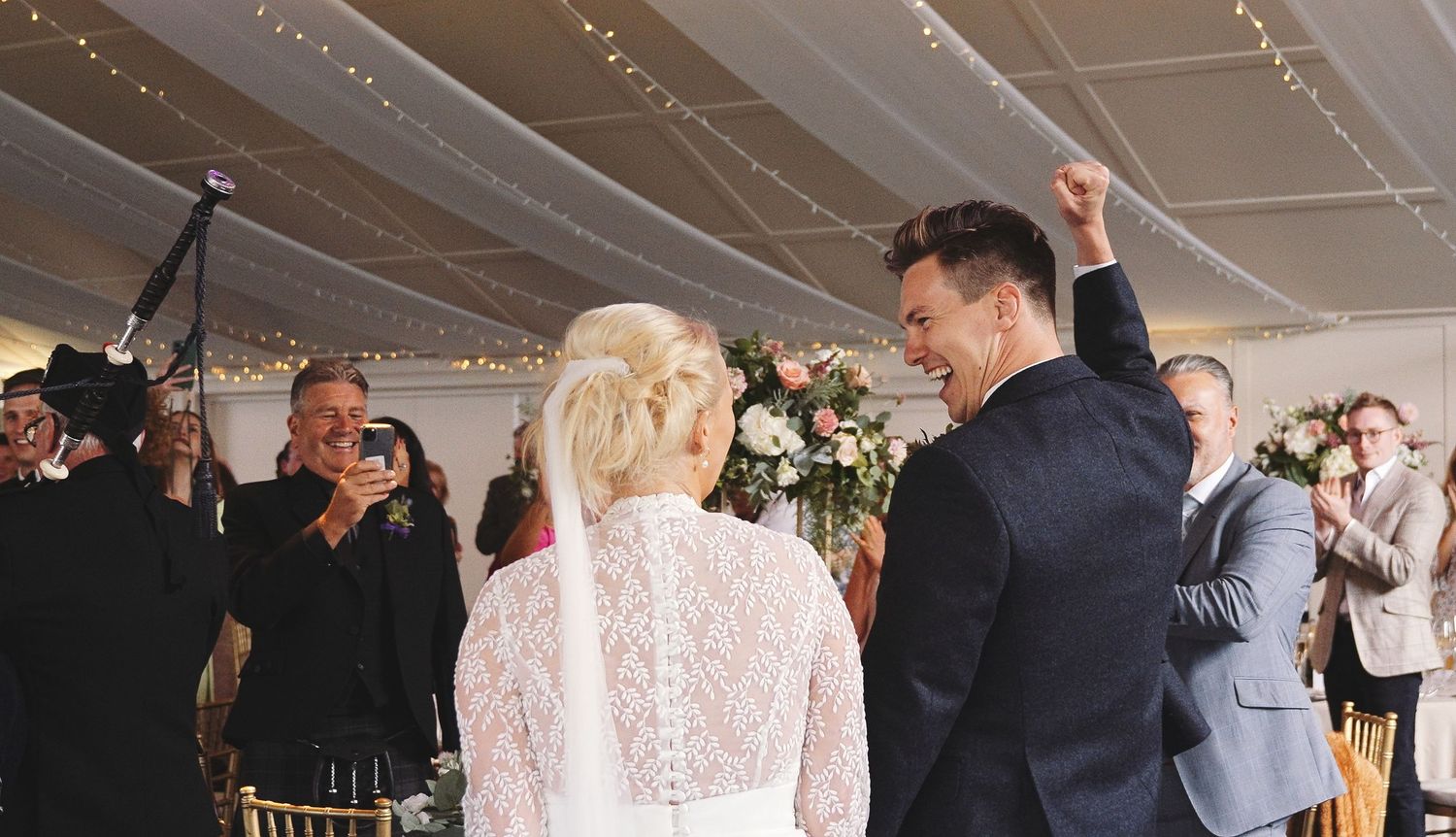 Joyful groom raises fist celebrating at wedding ceremony while bride in lace dress and veil stands before him.