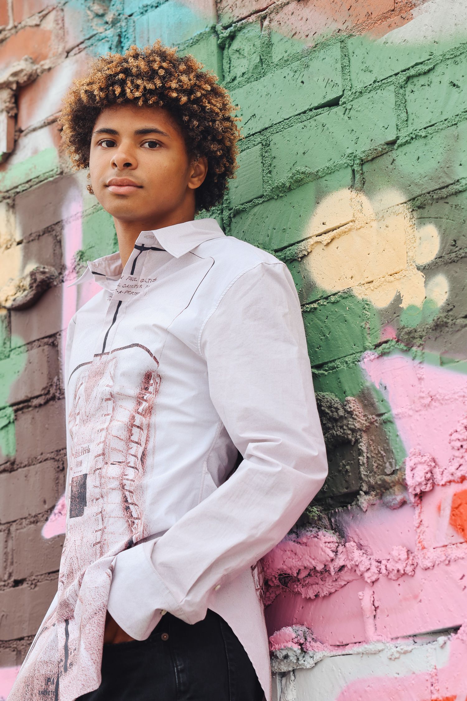 A stylish pose against a vibrant pastel painted brick wall wearing a white ruffled shirt.
