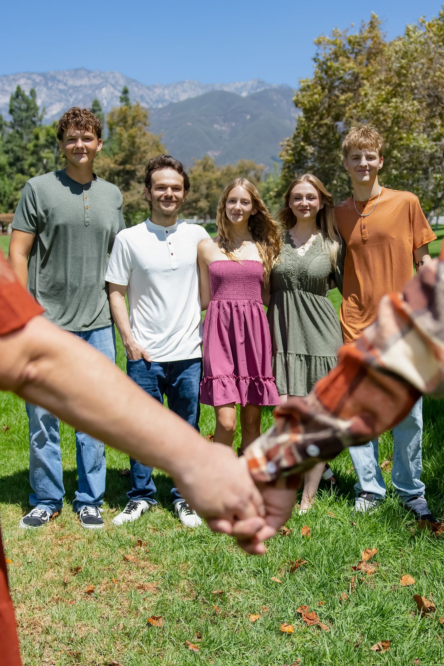 Group of five young adults smiling outdoors on a sunny day with mountains and trees in the background.